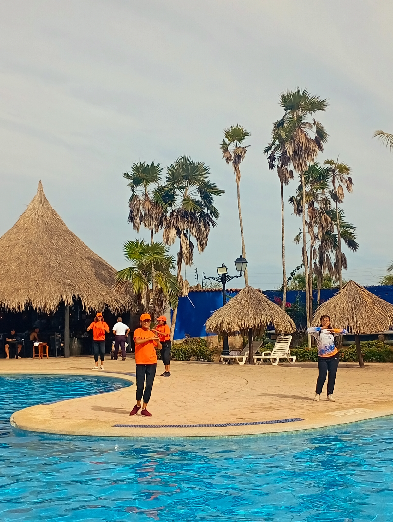 Scene: A daytime shot at a resort in Playa El Agua, Venezuela.
People: Several individuals are present. Three people dressed in orange t-shirts and dark pants are gathered on a concrete platform next to a pool. One of these individuals is wearing a hat. A fourth person stands alone, wearing a white and blue shirt with dark pants. A group of people sits under a large thatched-roof structure.
Objects: A blue-water pool, a large thatched-roof structure with people sitting beneath, smaller thatched-roof umbrellas, lounge chairs, palm trees, and street lights are visible.
Actions: People in orange shirts are standing and possibly exercising on the concrete platform next to the pool. The individual in the white and blue shirt is standing with arms outstretched. People are sitting beneath the large thatched-roof structure.
Environment: The scene appears to be a resort setting on a sunny day, with blue skies above. Vegetation includes palm trees.