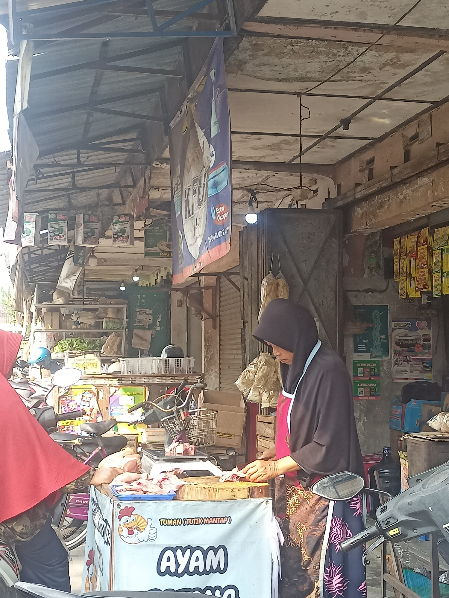 A woman wearing a black hijab and an apron is seen standing behind a market stall in Gemolong, Indonesia. She is actively engaged in chopping meat on a wooden cutting board. The stall itself is adorned with a banner that includes text in Indonesian, and features an illustration of a chicken giving a thumbs-up. Raw chicken meat is displayed on a blue tray on the stall. The stall is cluttered with various goods, including what appear to be packaged food items and bags of produce. Behind the woman, a metal door and shelves filled with merchandise are visible. The market environment appears to be outdoors, with a corrugated metal roof overhead supported by exposed beams. Several motorcycles are parked around the stall, suggesting a busy local marketplace. The overall scene depicts a typical day at a local food market.