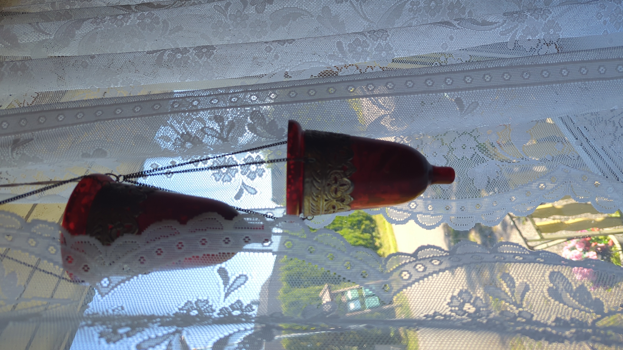 Two red glass candle holders hang by chains in front of a white lace curtain, obscuring a view of a residential area in Kløfta, Norway. The candle holders are decorated with intricate gold metal filigree around the rim. Through the lace and between the hanging candle holders, a blurry landscape of green trees, a house, a road, and a blue sky can be seen. The scene appears to be indoors, looking out of a window.