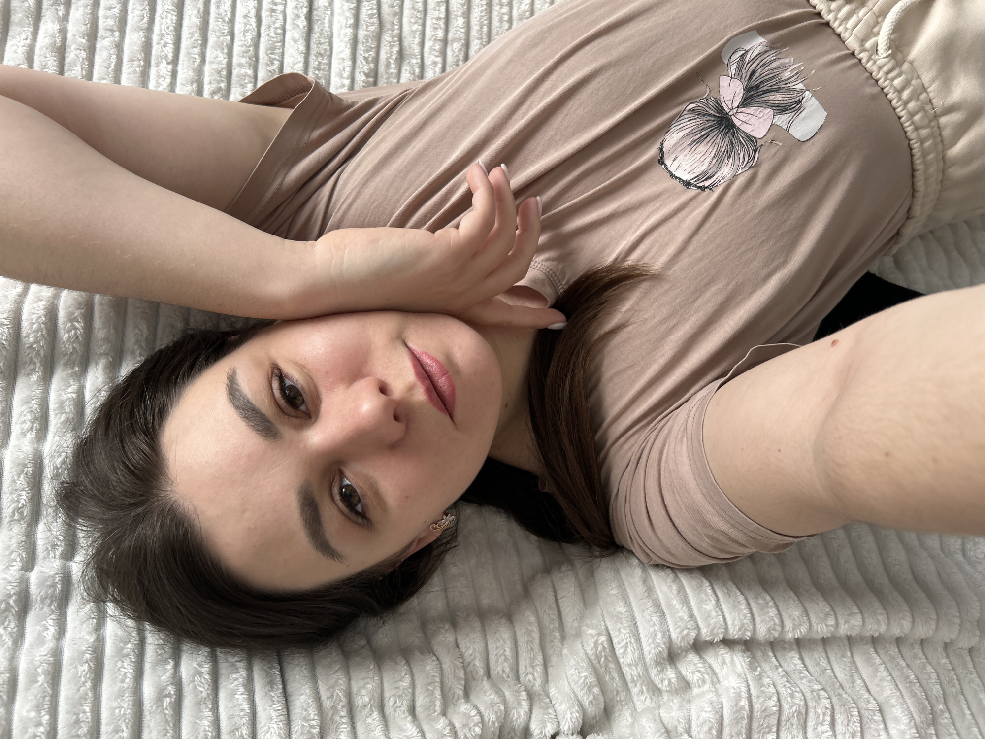 A young woman with brown hair and brown eyes is lying on a white, ridged blanket, possibly a bedspread. She is wearing a light brown t-shirt with a graphic of a hair bun on the front and light-colored shorts. Her head is tilted, and she is looking directly at the camera with a slight smile. Her left hand is gently placed near her face. She is wearing a small earring in her left ear. The background is simply the blanket. The location cannot be determined from the image.