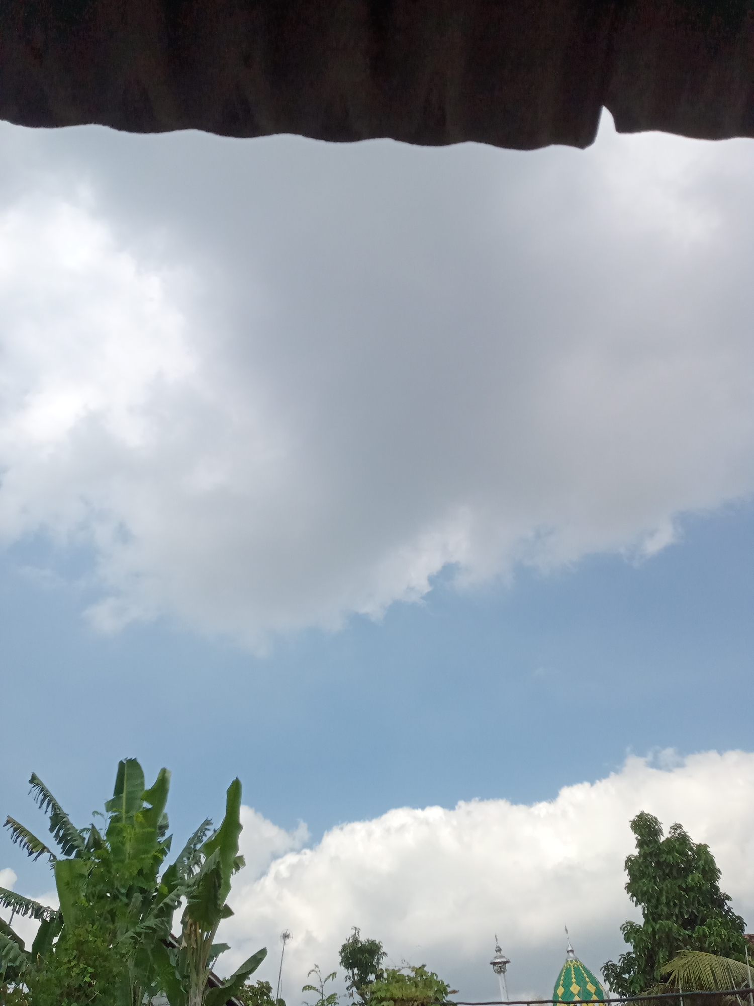 The image is a photograph taken from a low angle, looking upwards towards a cloudy sky. In the foreground, the dark, corrugated underside of a roof or overhang partially obscures the top of the frame. Below this, a vast expanse of sky dominates, filled with large, white cumulus clouds against a backdrop of pale blue.
The lower portion of the image reveals the tops of several trees, notably a lush green banana tree with large, tattered leaves on the left. To the right, other trees are visible, including a tall, dark green tree. In the distance, between the trees, parts of a building with a green, diamond-patterned dome and spires can be seen, suggesting it might be a mosque. A slender tower or minaret is also visible to the left of the domed building.
The scene appears to be an outdoor setting in a residential or semi-rural area, likely during the daytime given the presence of sunlight illuminating the clouds. The overall impression is one of a partly cloudy day.