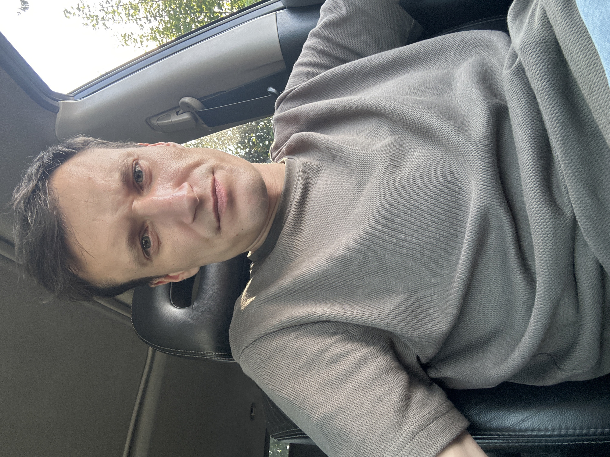 A man sits in the driver's seat of a car. He is wearing a grey t-shirt. The car's interior is visible, including the seat and the headrest. Sunlight streams in from the right side of the car, illuminating the trees and foliage outside. The image appears to have been taken during the day. The location in the city of Shatava, Ukraine, is not discernible from the image itself.