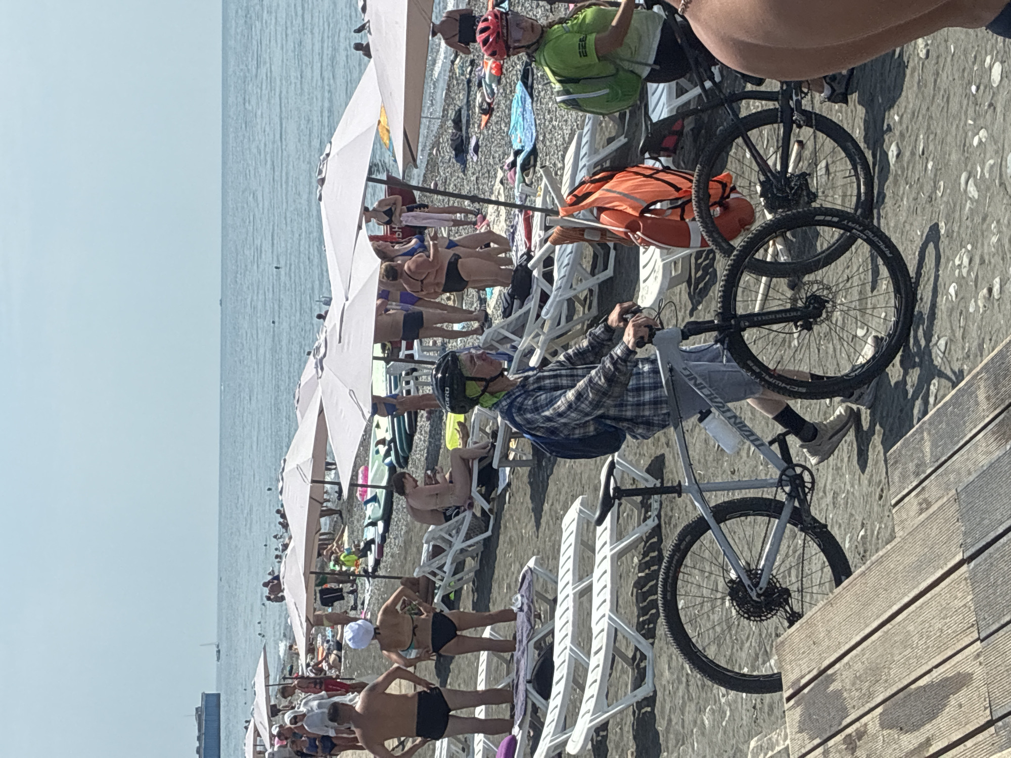 On a sunny day, a crowded beach in Sirius, Russia, is depicted with people relaxing and enjoying the coastline. The beach is covered in dark pebbles, and numerous lounge chairs and umbrellas are set up for visitors. The calm, blue sea stretches to the horizon under a clear sky.
In the foreground, two individuals are on bicycles. One person, wearing a plaid shirt and a green head covering, is on a gray bicycle, facing left. Another bicycle, black with red accents, is parked nearby, and a person in a green shirt and a red helmet is standing next to it, facing right. A bright orange life vest is hanging on a pole between them.
Further back, numerous beachgoers are visible. Several men and women are in swim attire, with some standing and others seated on lounge chairs. Umbrellas provide shade for various groups. In the background, a large, dark structure, possibly a ship or barge, is visible on the water, along with a small sailboat. The overall scene conveys a typical summer day at a popular beach destination.