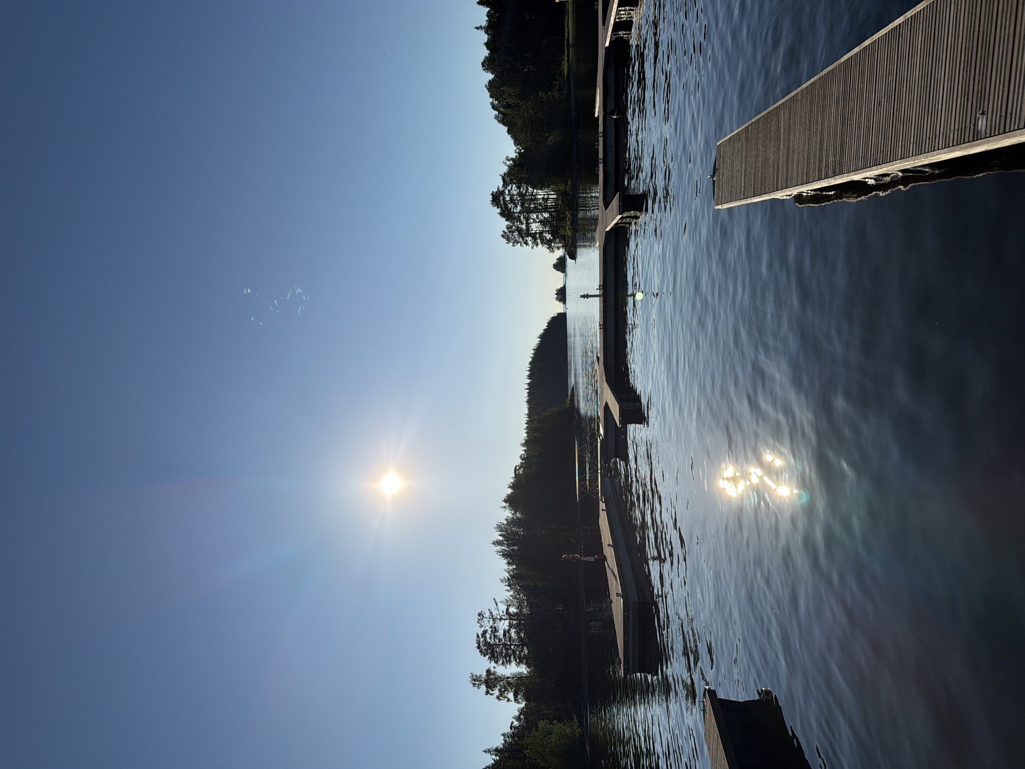 This is a vertical, eye-level, outdoor, full shot of a body of water with several wooden piers extending into it. The sun is shining brightly in the upper left portion of the image, casting a strong glare on the water and creating bright reflections. The sky is a clear, bright blue.
The water is calm with gentle ripples. The piers are constructed of wood and appear to be used for docking boats. One pier is in the foreground on the right, extending diagonally into the water. Another longer, rectangular pier is visible in the middle left of the image. Several other sections of piers are scattered across the water.
Along the edges of the water, there is dense, dark green forest. The trees are tall and appear to be pine or fir. The overall impression is of a peaceful, natural setting, likely a lake or bay. There are no buildings or prominent landmarks visible in the image. There are two small, silhouetted human figures visible on one of the piers in the middle distance, appearing to stand still.