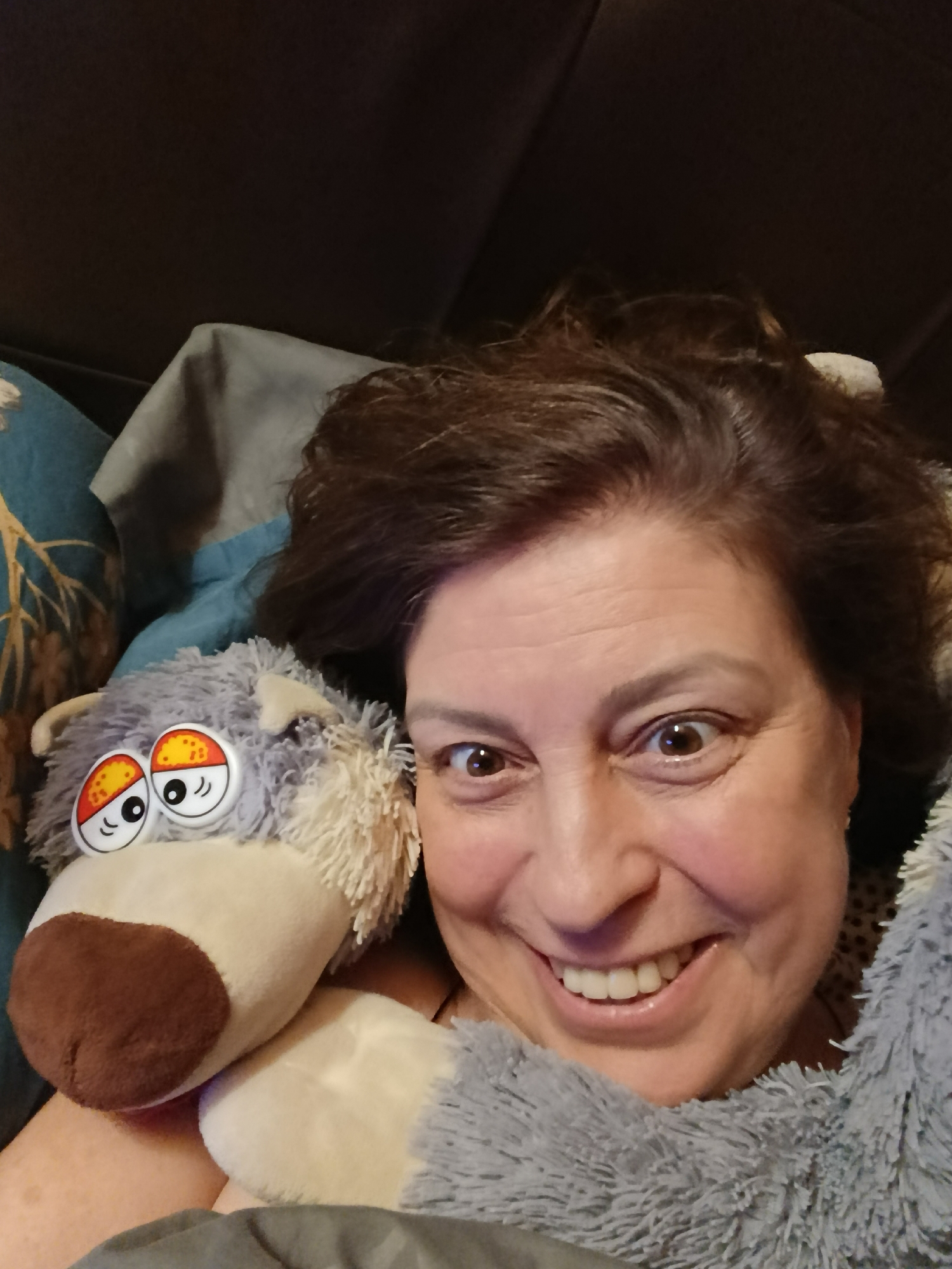 This close-up selfie, taken indoors in Verea, Russia, features a smiling woman alongside a plush toy. The woman, who appears to be middle-aged, has dark brown hair and a broad, joyful smile with her teeth visible. Her brown eyes are wide open, conveying an expression of surprise or delight. She is partially enveloped in a soft, fluffy grey blanket or garment, suggesting comfort and warmth.
To her left, a grey plush toy with a white muzzle and a brown nose is snuggled against her. The toy features large, exaggerated cartoon-style eyes with distinct red and yellow patterns on a white base, and shaggy lighter-colored fur around its face. It appears to be a dog or wolf character.
The background is dark and indistinct, indicating an indoor setting, likely a bed or couch, with soft furnishings. A patterned pillow with blue fabric and light-colored foliage designs is visible behind the woman's left shoulder and the plush toy. The lighting is consistent with an indoor environment, and the time of day is not explicitly discernable, but the cozy scene implies a relaxed, comfortable moment. No text is visible in the image. The overall mood is one of lightheartedness and contentment.