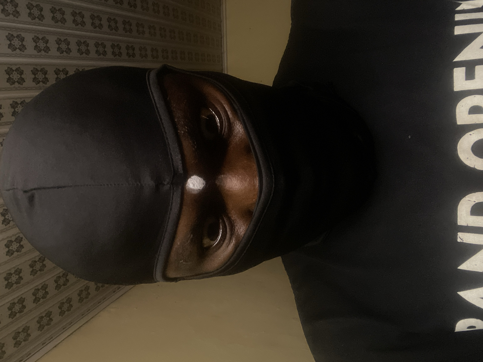 A person wearing a black balaclava is looking directly at the camera. A small, white, circular object is positioned on the balaclava between the person's eyes. The person is wearing a black hooded sweatshirt with partial, white, sans-serif text visible on the right side of the image, with the letters "AND OPENI" discernible. The background includes a section of patterned wallpaper, suggesting an indoor location. The image is taken in Jos, Nigeria.