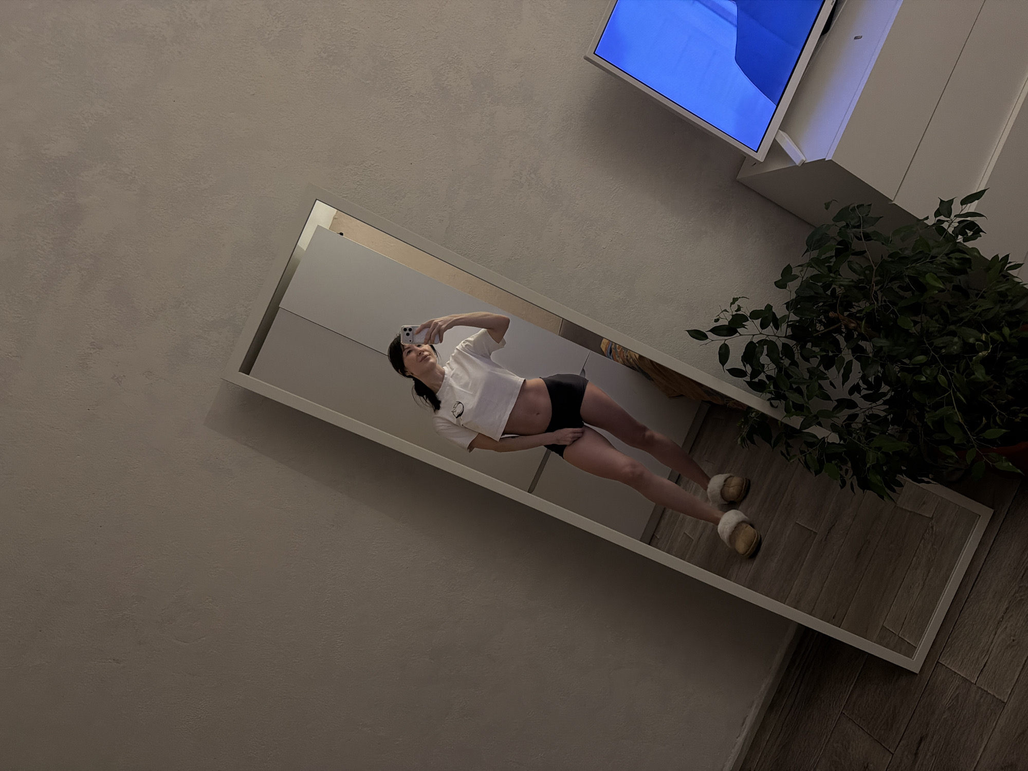 An indoor mirror selfie is presented from a tilted perspective, capturing a person in a room in Perm, Russia. A full-length, white-framed mirror occupies the lower part of the diagonal frame, reflecting the person taking the photo. The individual, appearing female, is holding a light-colored smartphone up for the picture. She is dressed in a white cropped t-shirt, dark shorts or underwear, and light-colored fluffy slippers. Her left hand is resting on her lower abdomen.
The wall behind the mirror is a textured light gray or beige. In the upper right section of the image, a television or monitor is mounted on the wall, displaying a bright blue screen. Below the television and extending partially behind the mirror is a piece of white, box-like furniture. To the right of the mirror, on a light-to-medium toned wood-look floor, a large potted plant with dark green leaves is visible. The scene is illuminated by artificial indoor lighting, suggesting it might be evening or night. No discernible text is visible.