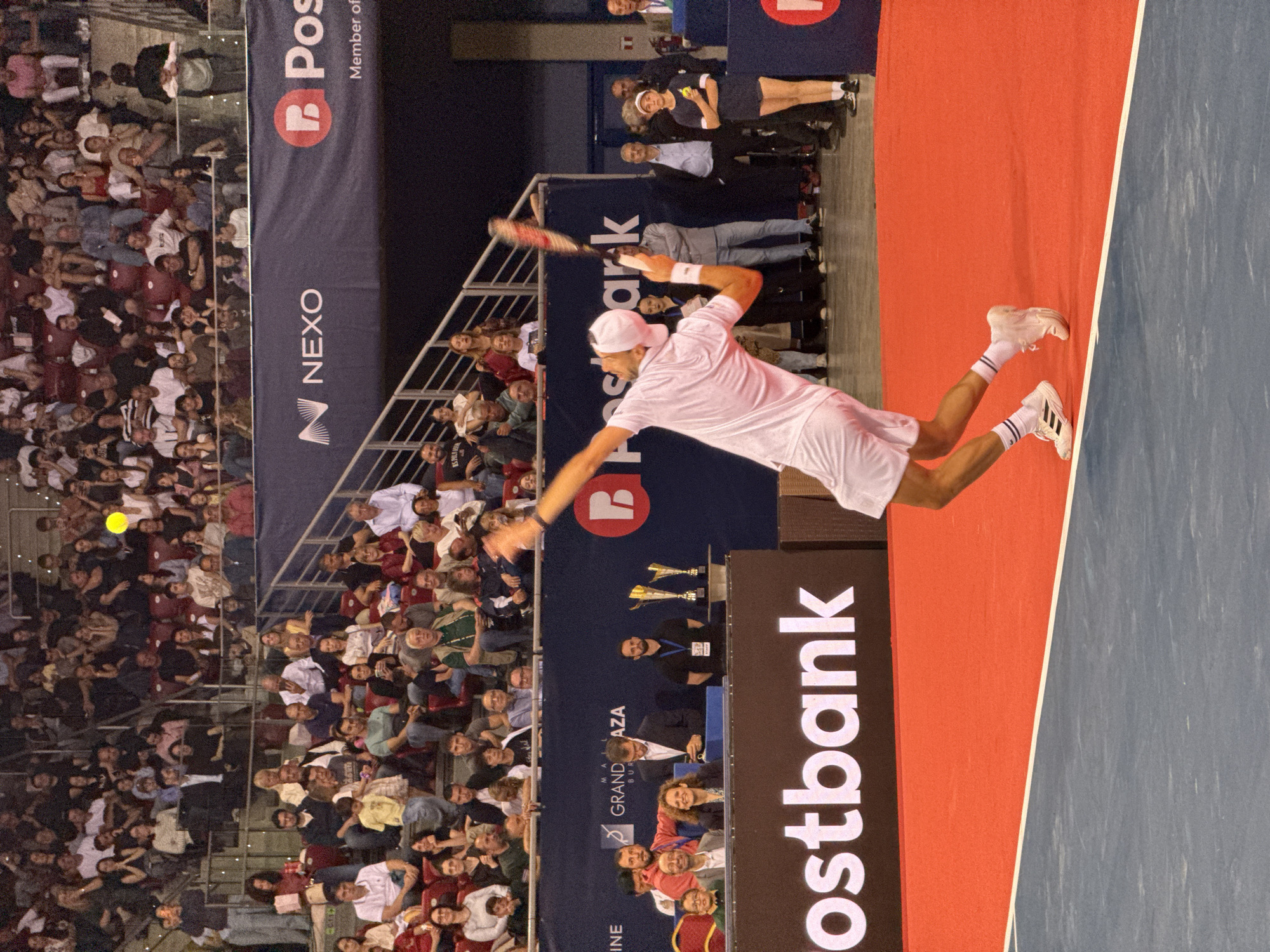 This image captures a tennis match, likely at the "Sofia Open," an ATP Tour event held annually in Sofia, Bulgaria. The primary subject is a male tennis player in mid-serve, wearing a white outfit, white cap, and white tennis shoes with dark stripes. He is captured in motion, with his body arched and racket raised, preparing to strike the ball, which is visible as a yellow orb in the air. The court features a red surface with a blue out-of-bounds area.
In the background, a large audience is seated in stadium seating, many of whom are watching the match. The stands are filled with people of diverse ages and attire. Behind the spectator area, banners are visible, including one that reads "NEXO" and another with a "P Postbank" logo, indicating commercial sponsorship of the event. Additional signs, such as "GRAND AZZZA," suggest the venue might be the Arena Armeec Sofia, a common location for such sporting events. Several officials and other individuals are positioned around the court and in the spectator area, including a young girl in a dark blue outfit and cap standing with her arms crossed, observing the game. The lighting suggests an indoor or evening event.