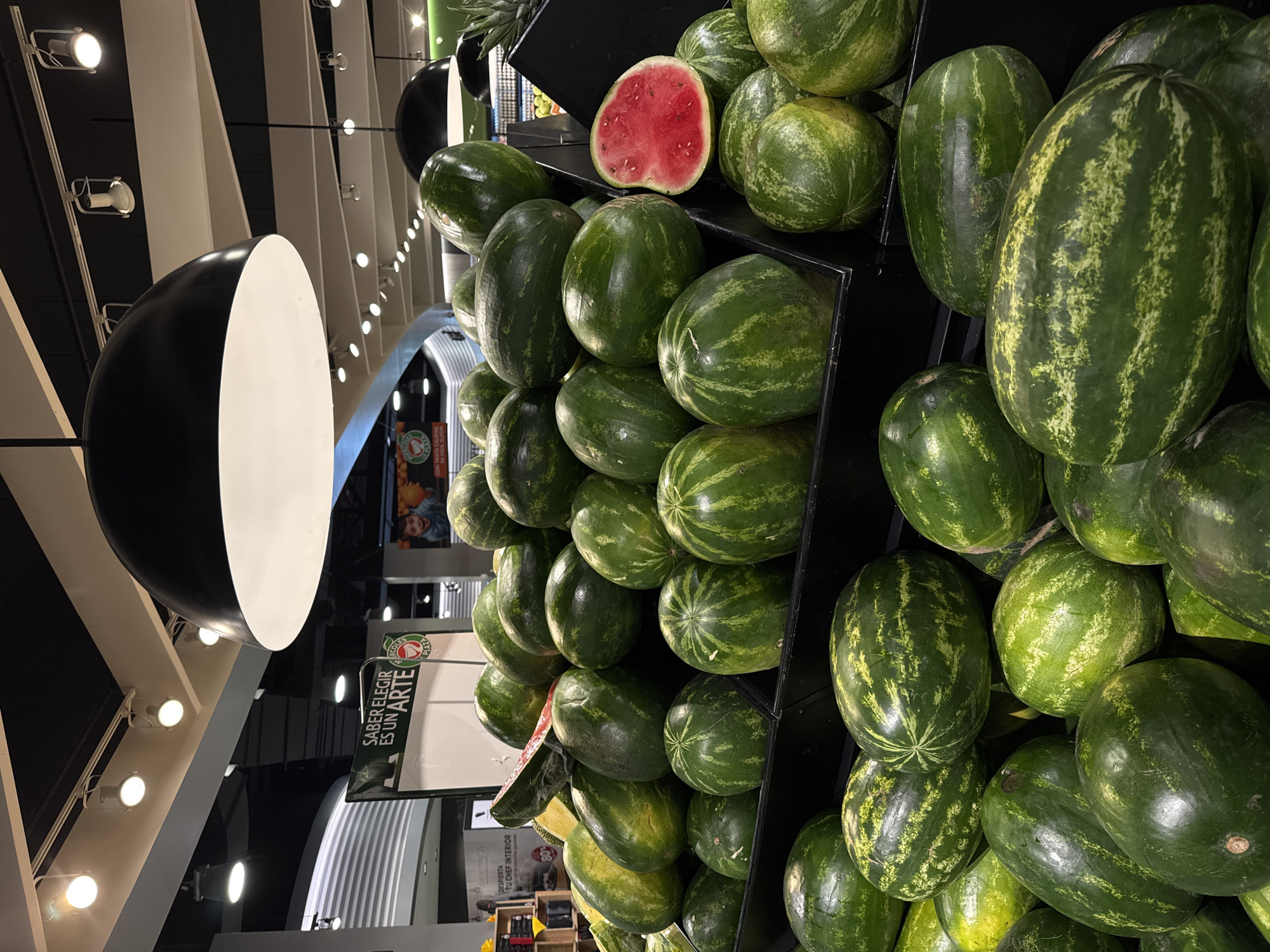 The image shows a display of watermelons in what appears to be a grocery store. The watermelons are green with dark green stripes and are stacked on black shelves. One of the watermelons has been cut open, revealing its red flesh. A black and white light fixture is visible in the background. A sign in Spanish that reads "SABER ELEGIR ES UN ARTE" (Knowing how to choose is an art) is partially visible. Based on the context, the location is likely in Puebla, Mexico, but this is not explicitly depicted. There are no people visible in the image. The focus is on the arrangement of watermelons in the store.