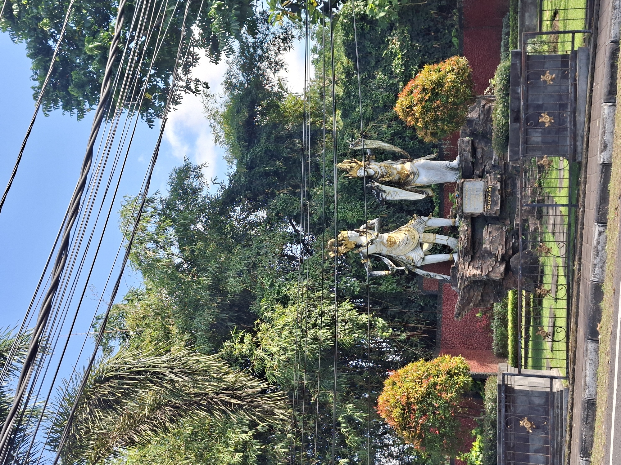 The image depicts an outdoor scene in Banjarangkan, Indonesia, with a prominent display of two ornate statues situated on a rocky base. These statues, appearing to be made of stone or a similar material, are adorned with gold accents and elaborate headdresses, each holding a distinct object or pose. One statue stands with a sword-like object, while the other has flowing drapery. Behind the statues, a lush green bamboo forest forms a dense backdrop, interspersed with other foliage. Several thick electrical wires are strung across the upper portion of the frame, silhouetted against a bright blue sky with scattered white clouds. In the foreground, a black metal fence with decorative gold floral motifs encloses a manicured green lawn. Two spherical, vibrantly colored bushes, one orange-red and the other with mixed green and red leaves, flank the statue display on either side. A section of a red textured wall is visible behind the bushes and to the left of the statues. The lower edge of the image shows a paved road and sidewalk, suggesting a location along a street.