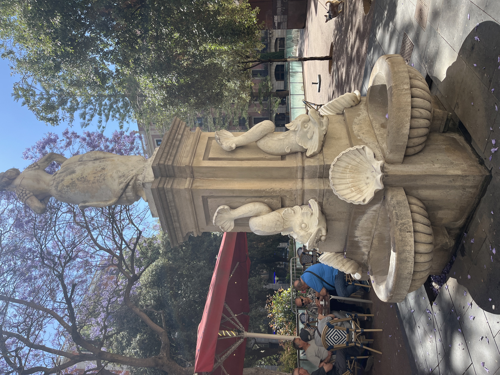 The image depicts a white stone fountain with decorative sculptures of sea creatures, including fish and seashells. The fountain is located outdoors in a public area. People are seated at tables nearby, under a red umbrella, suggesting a cafe or restaurant setting. In the background, there are buildings and trees, some with purple blossoms. The fountain appears to be a historical monument or public art installation. The location is likely Barcelona, Spain, given the architecture and the presence of jacaranda trees.