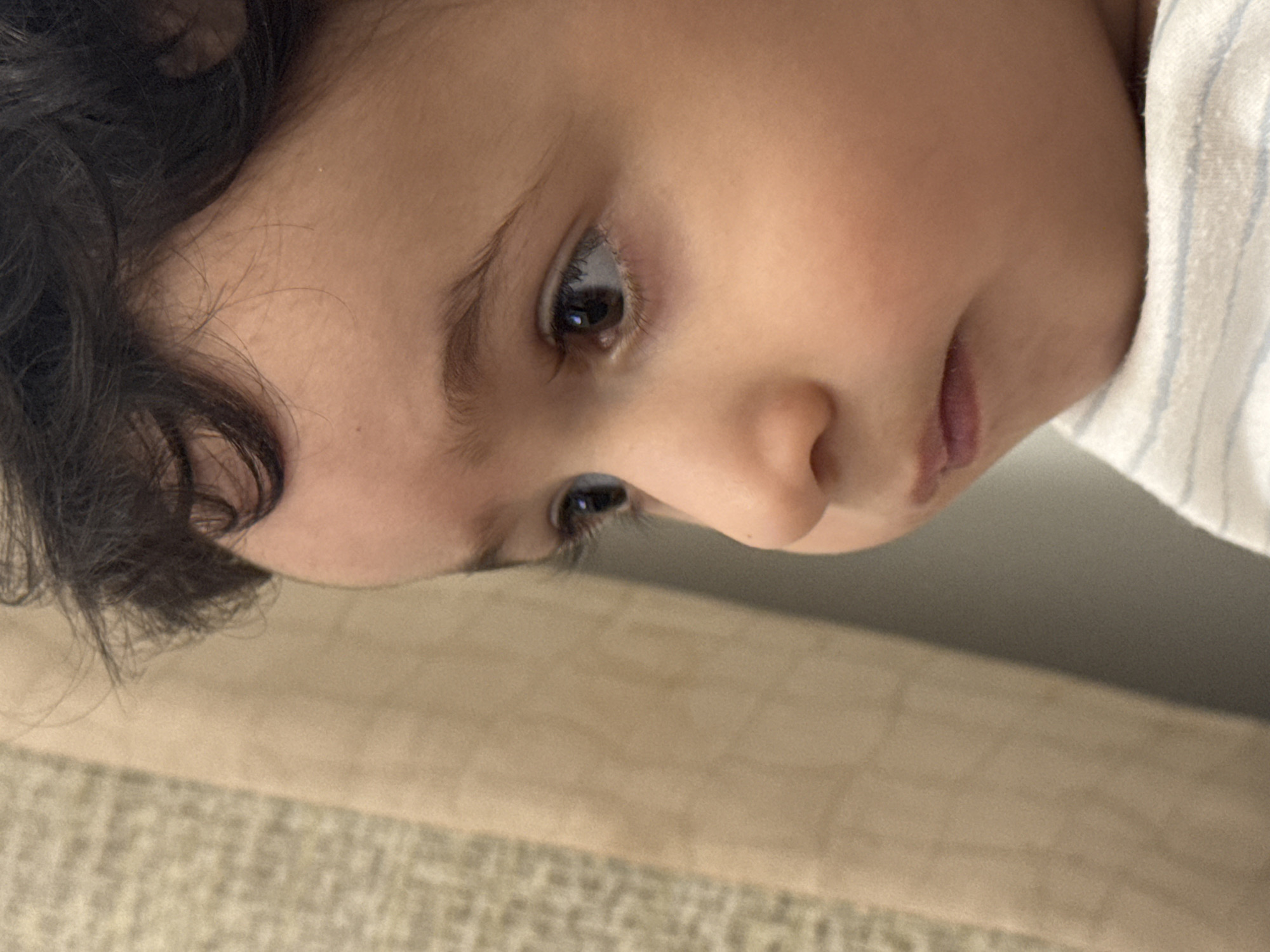 A close-up, profile shot focuses on the right side of a young child's face, with the child looking to the right. The child has dark, curly hair and a light complexion. Their right eye is visible, dark brown with long eyelashes. The child's nose and mouth are also visible, with the lips slightly parted. The child is wearing a white shirt with thin blue stripes. In the background, to the left of the child, is a beige, textured curtain or fabric. The lighting suggests an indoor environment, possibly near a window. The image is cropped tightly, showing only the upper chest and face of the child. There is no indication of the city of Sabunchu Raion, Azerbaijan, or any urban elements.