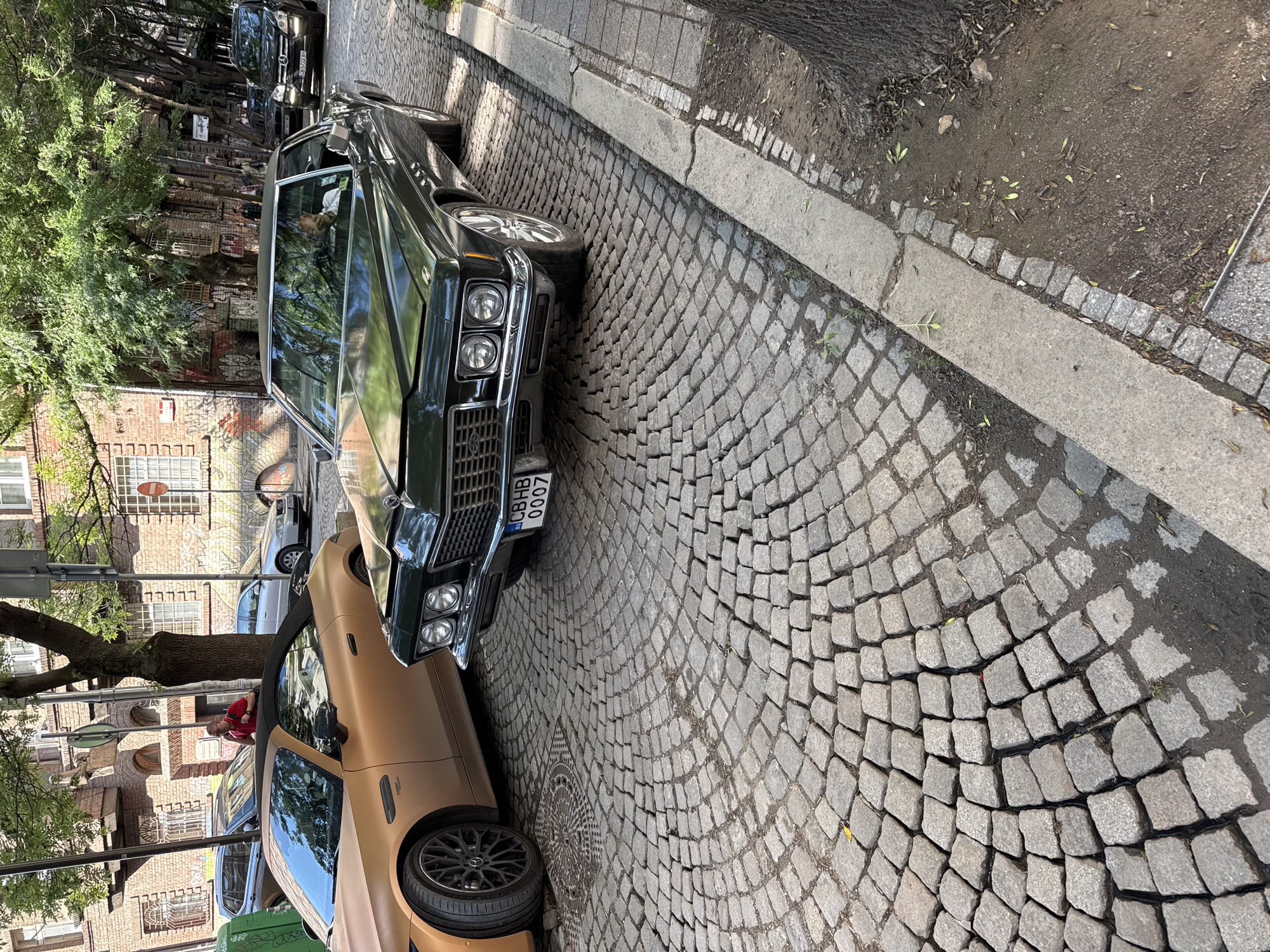 The image shows a street scene in Sofia, Bulgaria. The street is paved with cobblestones and lined with parked cars. Two cars are in the foreground. The first car is matte gold with custom wheels. The second car is dark green and appears to be a vintage vehicle with a "CB HB 0007" license plate. A man is leaning into the matte gold car. Other parked cars and trees are visible in the background, and a brick building is across the street.