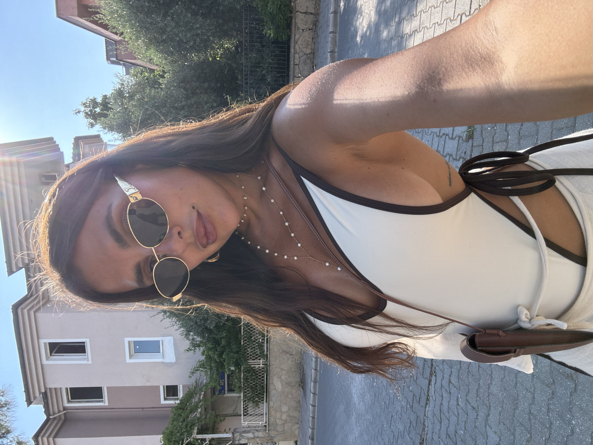 A woman is taking a selfie outdoors. She is wearing a white swimsuit with dark trim and a small brown purse hanging from her shoulder. She has on oval-shaped sunglasses and a pearl necklace. The background shows buildings, trees, and a paved street. The scene appears to be in a residential area. The sunlight suggests it is daytime.