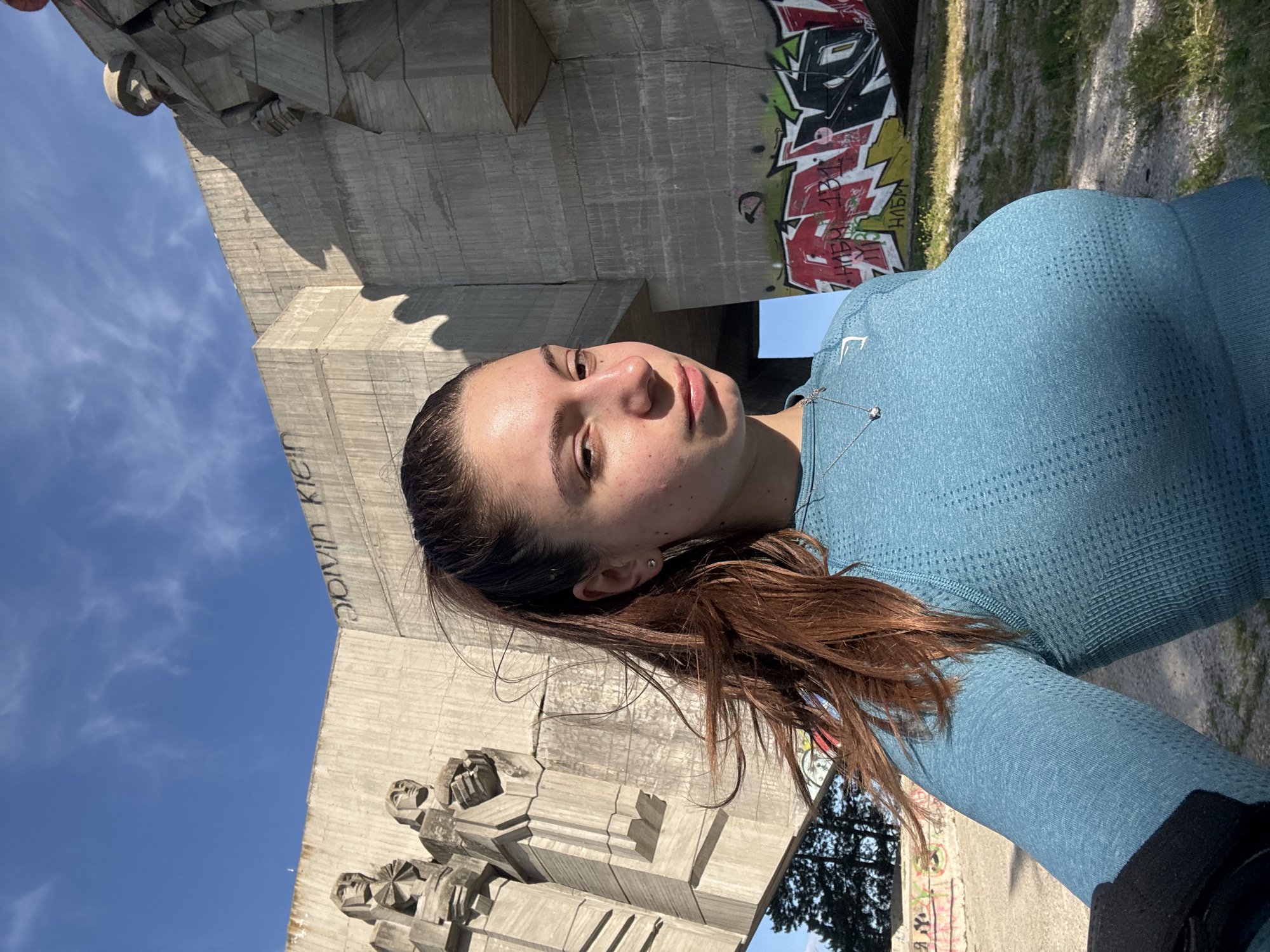 A young woman with brown hair and a light blue athletic top is taking a selfie outdoors. She appears to be in Varna, Bulgaria, standing in front of the Monument of the Bulgarian-Soviet Friendship. The monument is a large concrete structure, and there is graffiti on the concrete behind the woman. The sky is blue and there are trees and grass visible in the background. The woman is wearing a necklace with a pendant. Her hair is loose and falls around her face and shoulders. She is looking directly at the camera with a neutral expression.