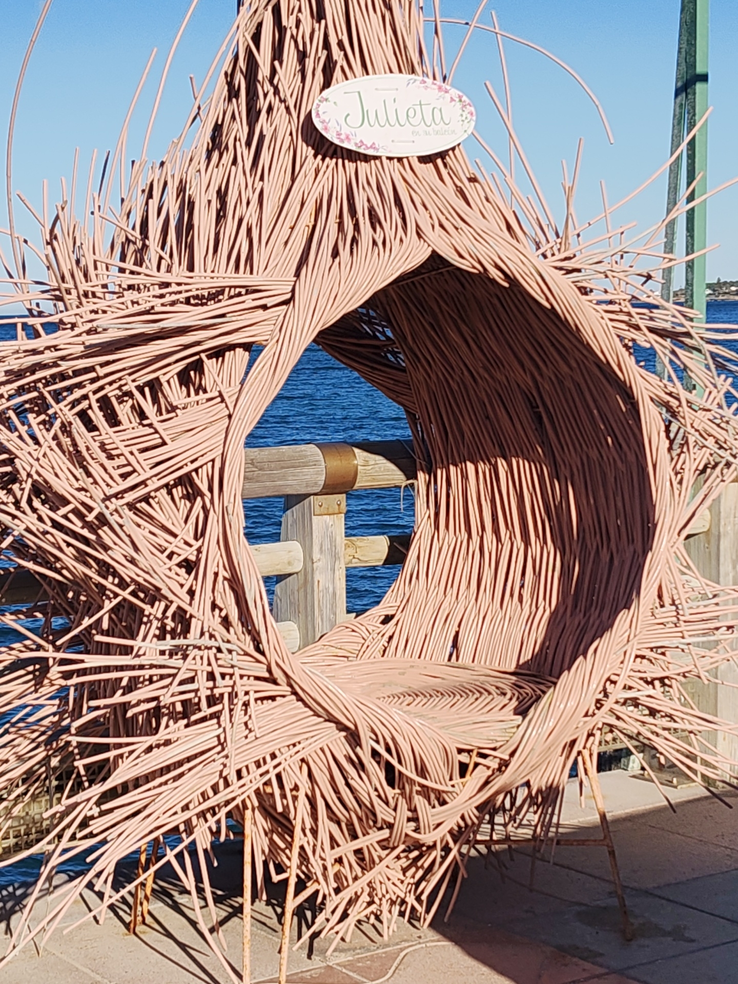A large, artistic chair, resembling a nest or cocoon, dominates the frame. It is constructed from numerous thin, woven strands of light brown or pinkish material, possibly wicker or branches. The chair features a prominent circular opening at its front, leading into a curved seating area. Many loose strands extend outwards from its body, creating a textured, somewhat spiky silhouette.
Attached to the top of the chair is a small, oval-shaped sign with a white background, decorated with green text and subtle floral patterns. The visible text clearly reads "Julieta," with smaller, less distinct text below it.
The chair is placed outdoors on a light-colored paved surface, likely a promenade or pier. In the background, a clear, brilliant blue sky meets a vast expanse of deep blue sea, indicating a coastal setting typical of Dénia, Spain. A sturdy wooden railing with metal fasteners runs horizontally behind the chair, separating the seating area from the water. Distant land or structures are faintly visible across the water.
The scene is bathed in bright natural sunlight, evidenced by the crisp, elongated shadows cast by the chair onto the pavement, suggesting a clear day during the late morning or early afternoon. The chair appears to be a decorative installation or unique seating for public use, perhaps for leisure or photography, with no individuals present in the image.