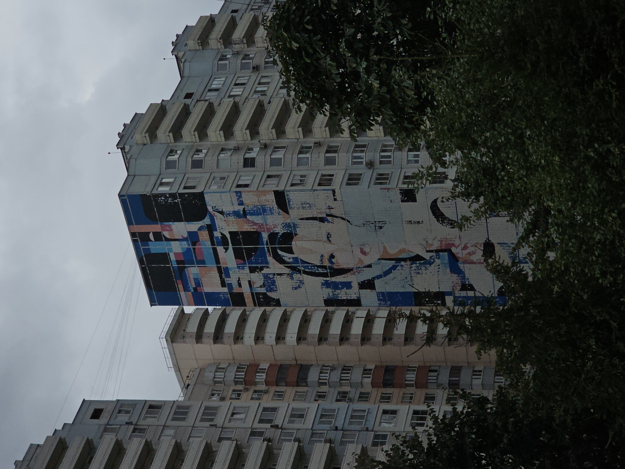The image shows a mural of a woman's face painted on the side of a tall, multi-story apartment building in Odintsovo, Russia. The building is light blue with many balconies and windows. The mural is abstract and colorful, with geometric shapes and a stylized portrait. Other apartment buildings are visible in the background, as well as some trees and a cloudy sky. The mural is a prominent feature of the building, and it is likely a work of street art.