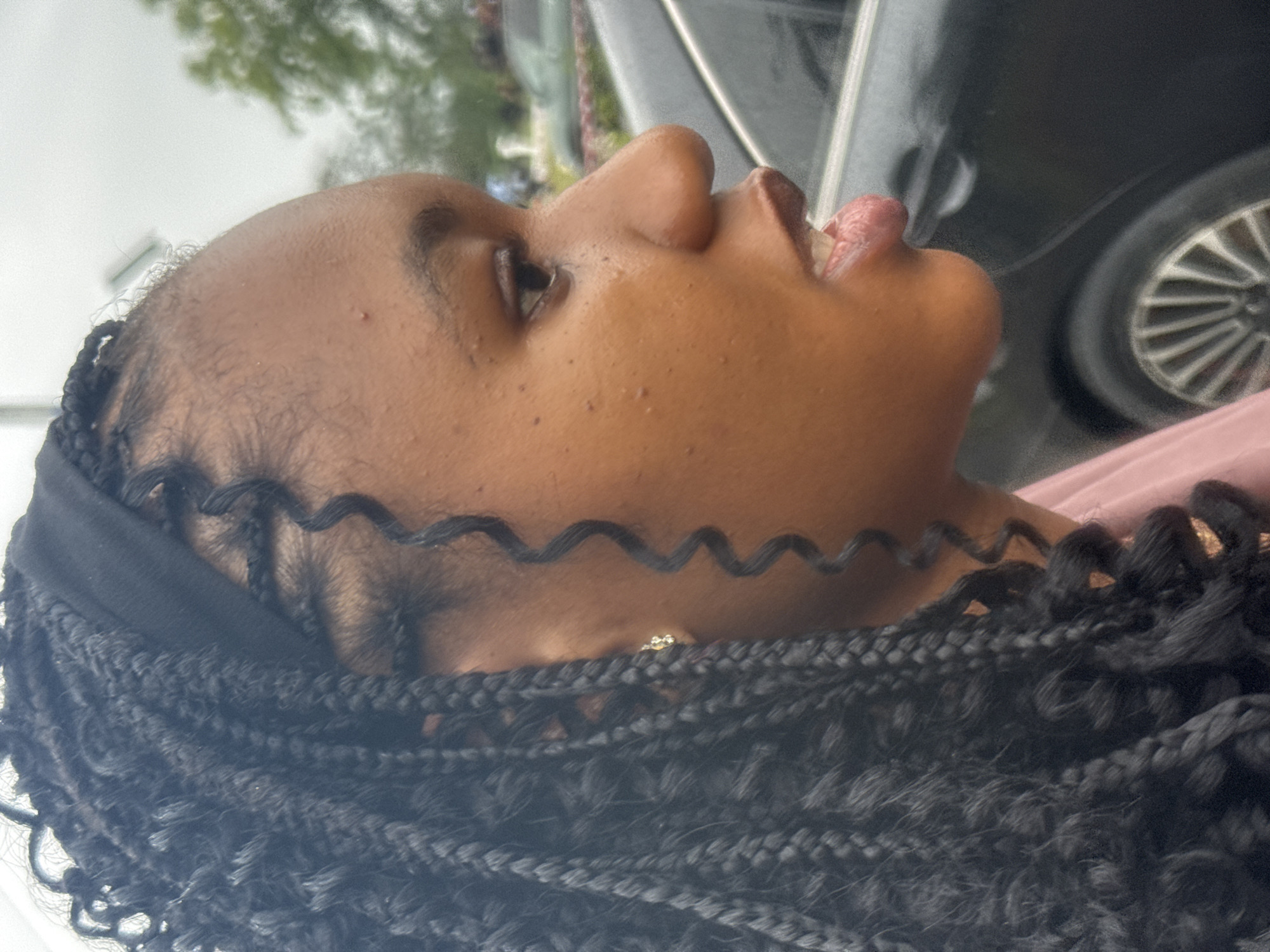 A young Black female is depicted in a close-up profile view, outdoors in Ihiagwa, Nigeria. She has a medium-dark skin tone and her hair is styled in long, dark box braids, some of which are curly or wavy. A black fabric headband is worn across her forehead. A small, light-colored, possibly diamond, stud earring is visible in her right earlobe. Her face shows several small, raised blemishes on her forehead and cheek. Her mouth is slightly open, revealing a portion of her lower teeth and the tip of her tongue, indicating a moment of speech or exhalation. A light-colored, possibly pink, garment is visible on her right shoulder. The background is significantly blurred, featuring a dark-colored sedan with silver-spoked wheels on the lower right, and indistinct green foliage against a light, overcast sky above it. No other individuals are visible in the frame.