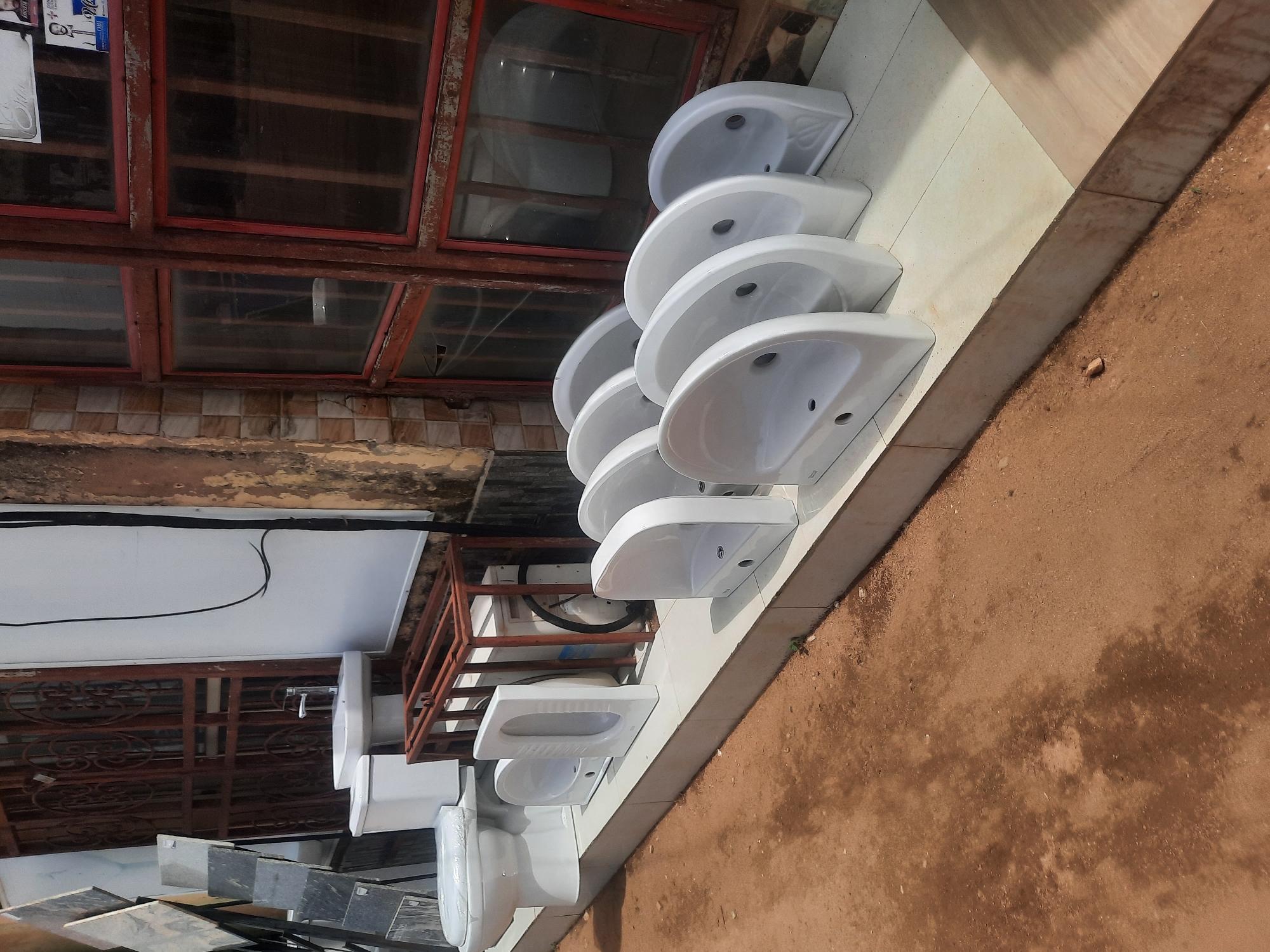 The image shows a collection of white ceramic sinks stacked along the exterior wall of a building in Gura Top, Nigeria. The sinks are positioned vertically, appearing to be for sale or storage. Adjacent to the sinks are stacks of what seem to be stone or tile slabs. The building's exterior features a window with a red frame and aged walls. The ground near the wall is dirt. Overhead, there is a white ceiling or overhang with visible electrical wiring.