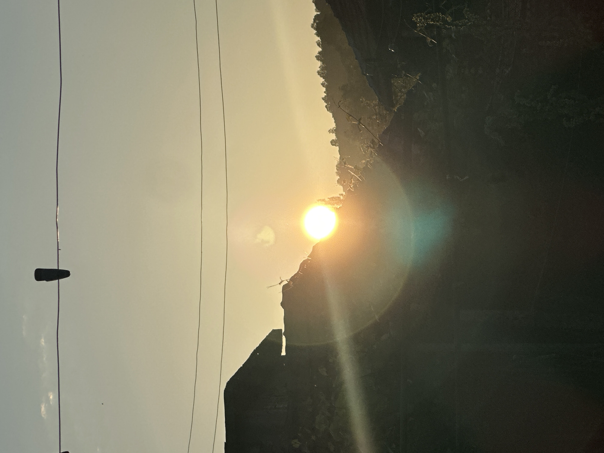 The image captures a vibrant sunrise or sunset over a silhouetted landscape. The sky, occupying the left two-thirds of the frame, transitions from a pale blue-gray on the far left to a warm yellow and orange hue towards the right. A brilliant, glowing sun is prominently featured on the right side, radiating strong light and a distinct diagonal lens flare extending downwards and to the right.
Three dark, thin lines, likely utility wires, run vertically from the top to the bottom of the image across the sky. A small, dark, clip-like object is attached to the leftmost wire in the upper portion.
The bottom right quadrant features a dark, silhouetted landscape, consisting of what appears to be hills or mountains. Sparse vegetation is visible along the crests of these dark forms. Beneath the sun, an angular, dark structure, possibly a building or ancient ruins, is visible in silhouette, merging with the landscape.
The overall scene suggests either early morning or late evening in a rural or mountainous area, likely near Slavyani, Bulgaria, given the context. The weather appears clear, with atmospheric conditions creating the soft, warm light and lens effects. No visible text is present in the image.