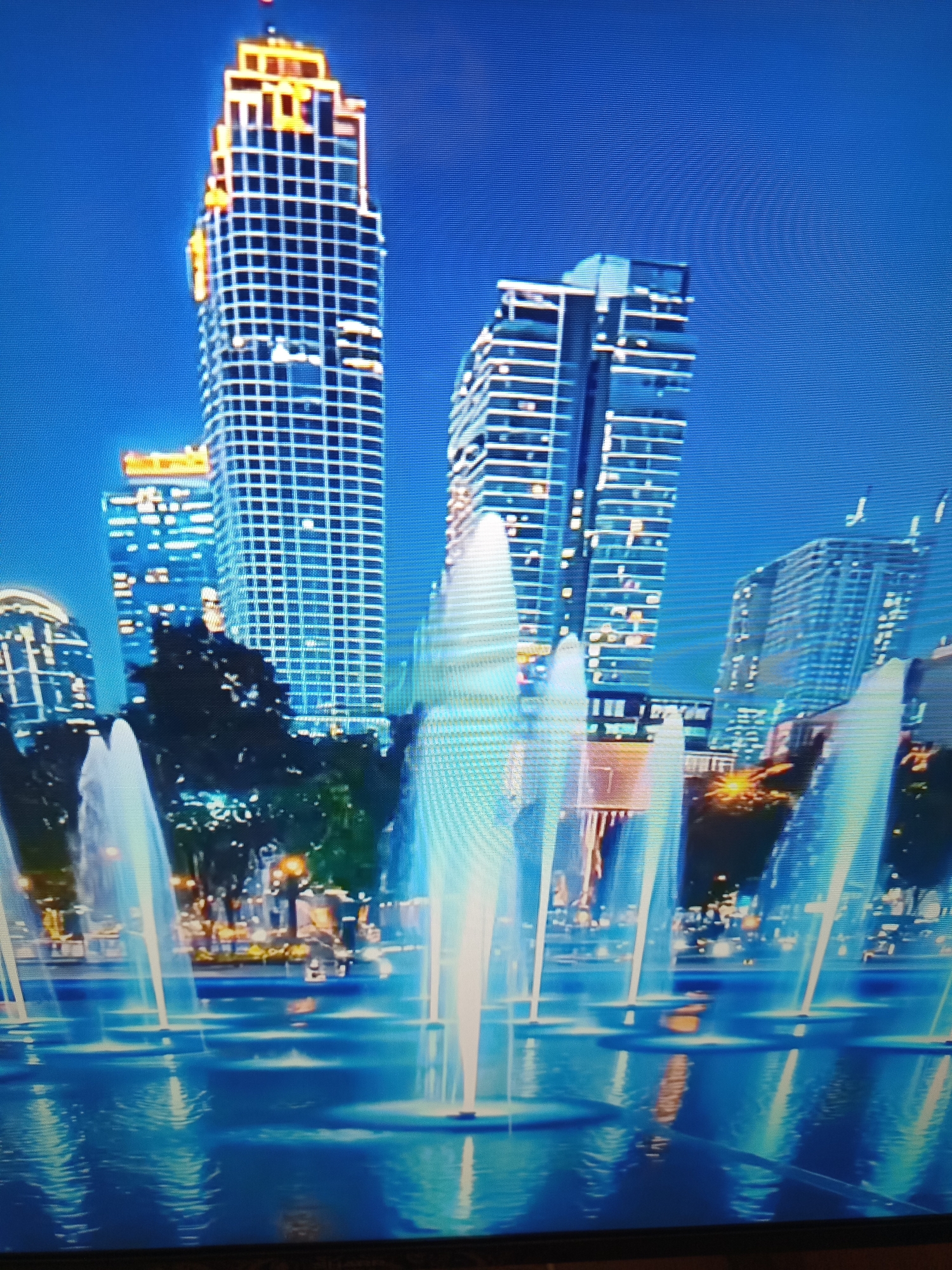 The image shows a nighttime view of a city skyline featuring several modern skyscrapers. In the foreground, a water fountain displays illuminated jets of water, creating a shimmering effect on the surface of the pool. The buildings in the background are lit up, with prominent examples including a tall, grid-like skyscraper with orange and white lighting near the top, and other buildings with horizontal white lighting elements. The sky is a deep blue. The scene is likely a public plaza or waterfront area in a metropolitan setting. There are no people or specific landmarks visible that would identify the city as Brno, Czechia.