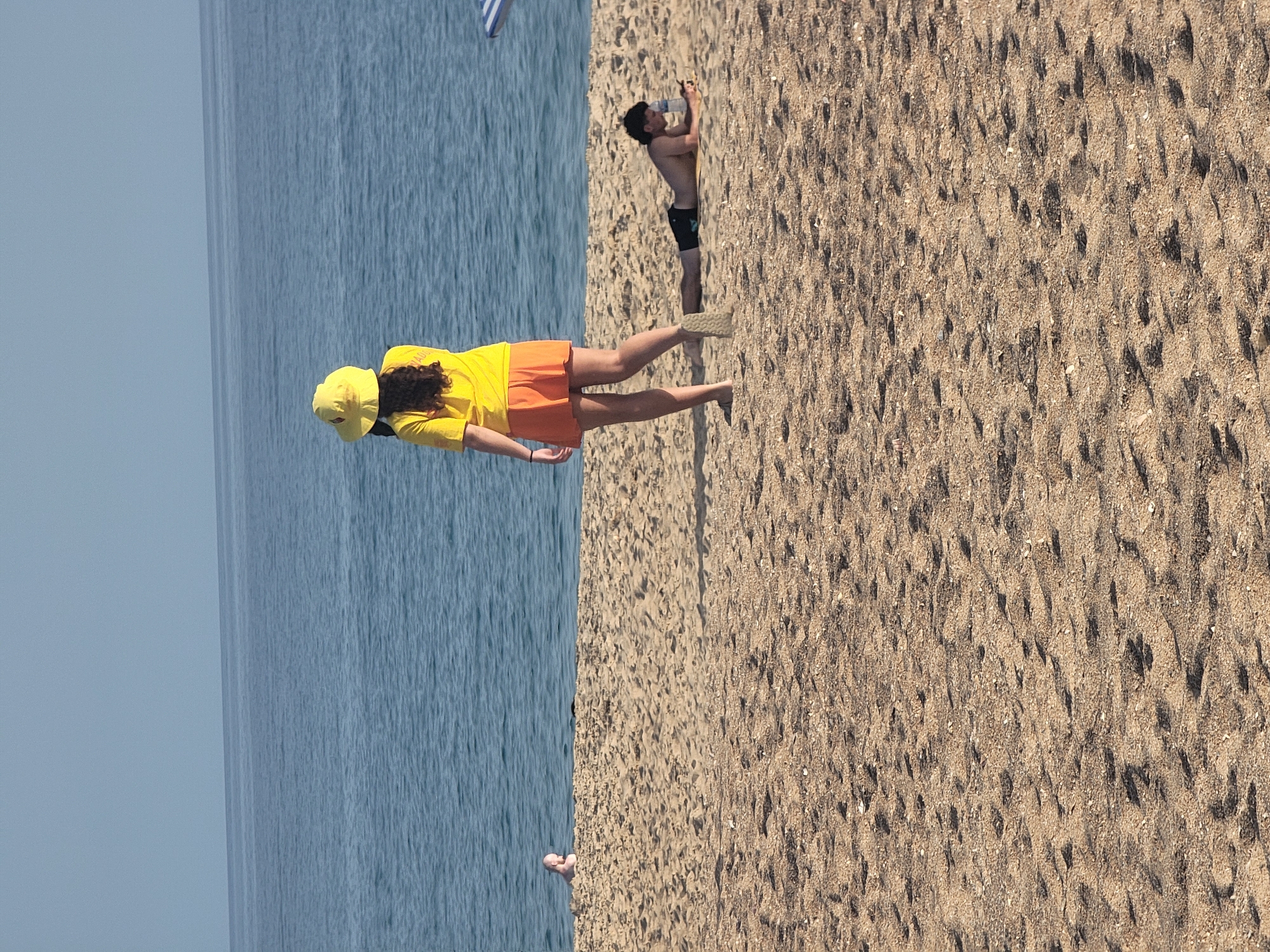 A medium shot captures a beach scene in Quarteira, Portugal, with the calm, blue Atlantic Ocean in the background and a sandy beach in the foreground. Two individuals are visible on the sand. In the middle ground, a person wearing a yellow sun hat and an orange skirt stands facing the ocean. To the right, a shirtless person wearing black swim trunks lies on the sand, looking at a phone and holding a water bottle. In the far background, a child is partially visible sitting in the water. The sand is covered with small shells and has footprints.
