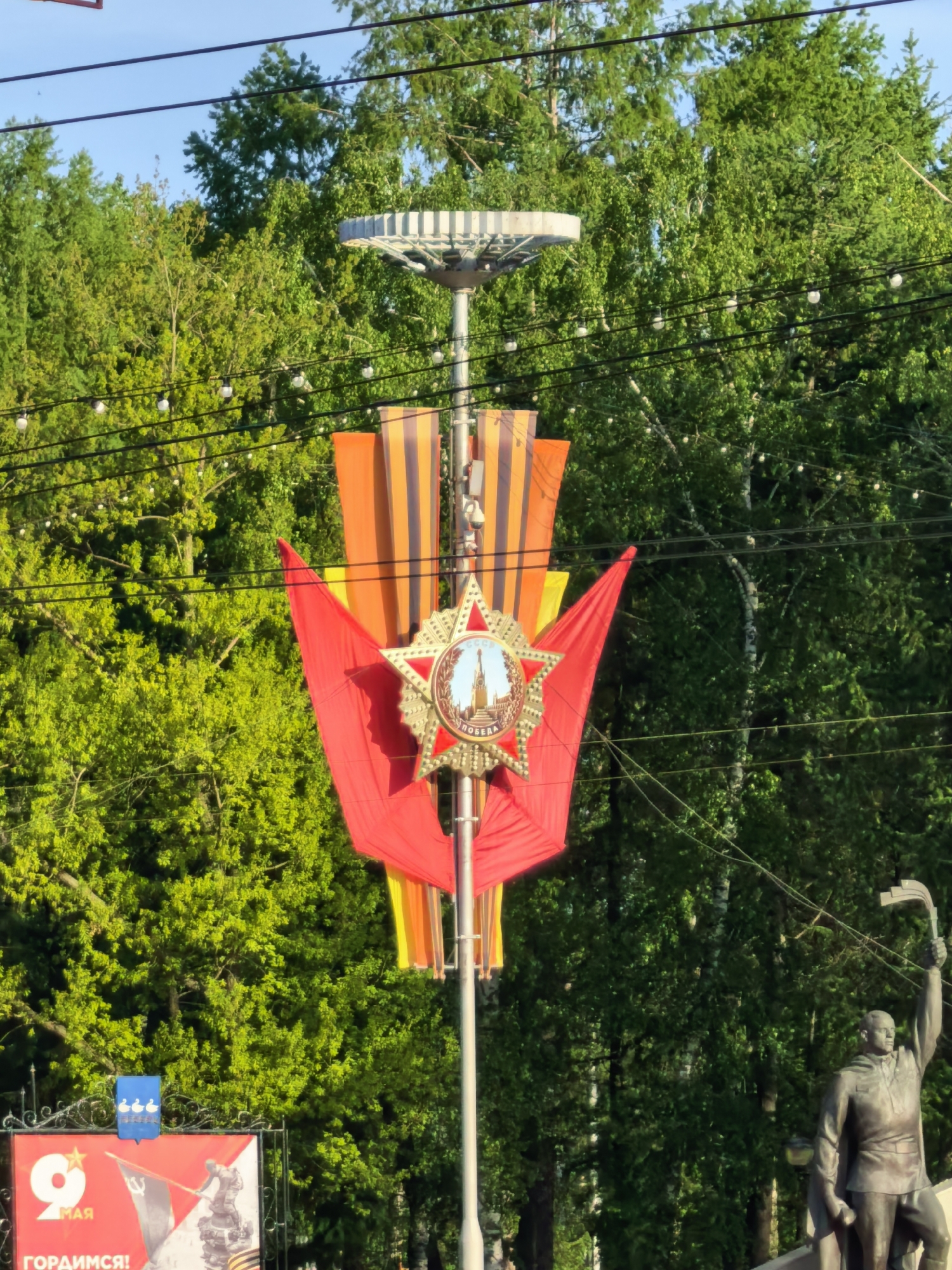 The image depicts an urban setting in Sterlitamak, Russia, likely a street decoration for a commemorative event. A light pole is adorned with a red flag, a Saint George ribbon, and a star-shaped medallion featuring an image of a monument, possibly within Moscow. In the background, lush green trees dominate, interspersed with power lines carrying strings of light bulbs. To the right, a statue of a man is visible, appearing to hold an object aloft. In the lower left, a billboard displays a depiction of a monument, as well as the Russian word for “May”.