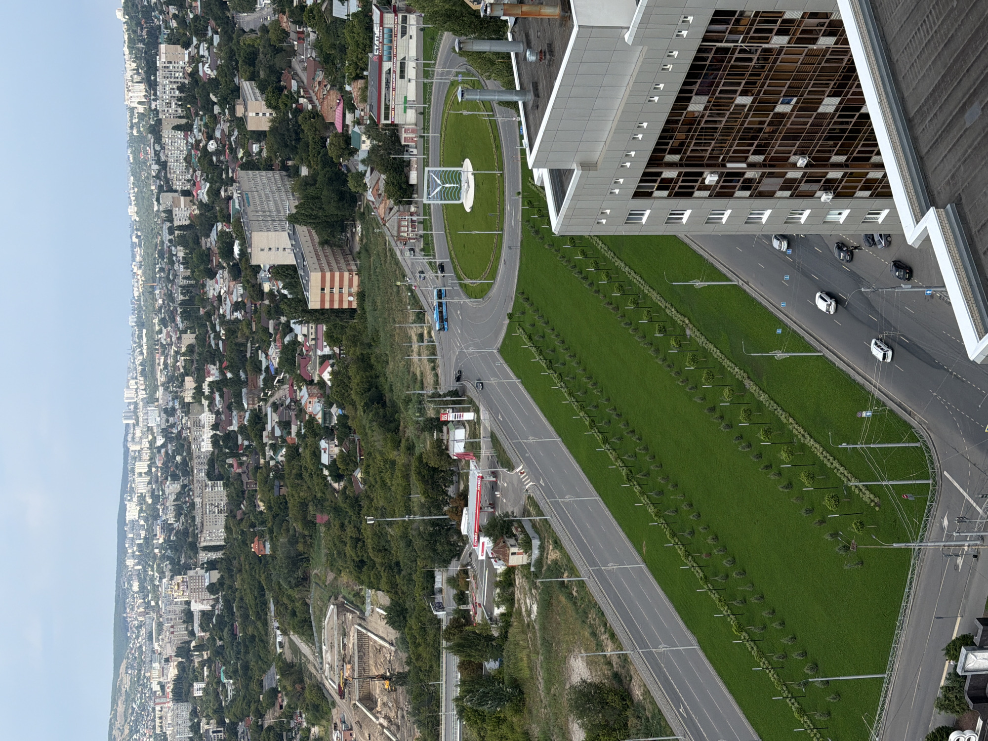 An aerial, high-angle, long shot shows a cityscape in Saratov, Russia. In the foreground, a multi-story office building with a glass facade and a dark roof juts into view. The building has many windows, some tinted brown and others reflecting the sky. In the middle ground, a wide, paved road curves around a circular grassy median with a modern sculpture in its center. A blue bus and several cars travel on the road. Adjacent to the road is a well-maintained green lawn with rows of young trees and lampposts. A gas station with red and white signage is visible to the left of the road. In the background, residential buildings, a construction site with earth moving equipment, and lush green trees dominate the landscape. Hills and a hazy sky complete the scene.