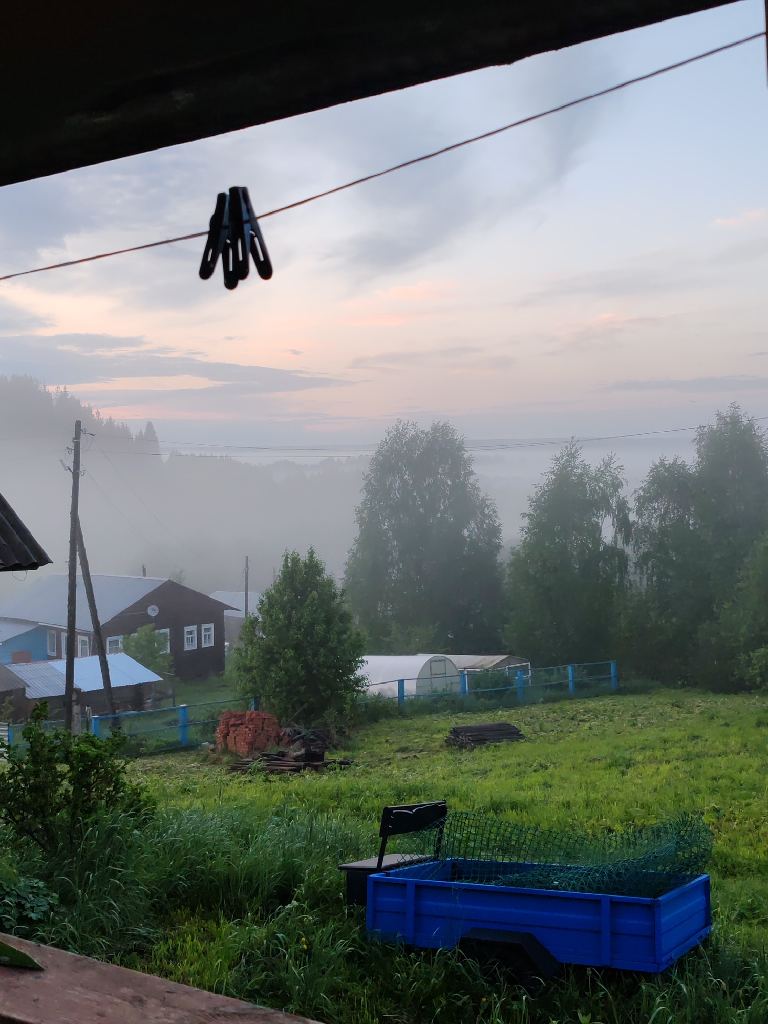 The image depicts a rural scene in Чухлэм, Russia, on a foggy morning. The view is from under an overhang, likely a porch or veranda, framed by dark beams. A clothesline with several black clothespins is strung across the foreground. Beyond the line, the scene opens to a yard with tall green grass. A small blue trailer with a mesh cargo restraint is visible in the lower right corner. A white greenhouse structure and a blue fence run across the middle ground. Beyond these, several houses with blue and brown siding are partially obscured by fog. The fog hangs heavy in the background, obscuring trees and creating a soft, diffused light in the sky, which shows tinges of pink and grey. A powerline or telephone line is visible cutting through the fog. A stack of bricks is beside a pile of dark materials. A wooden fence is to the left.