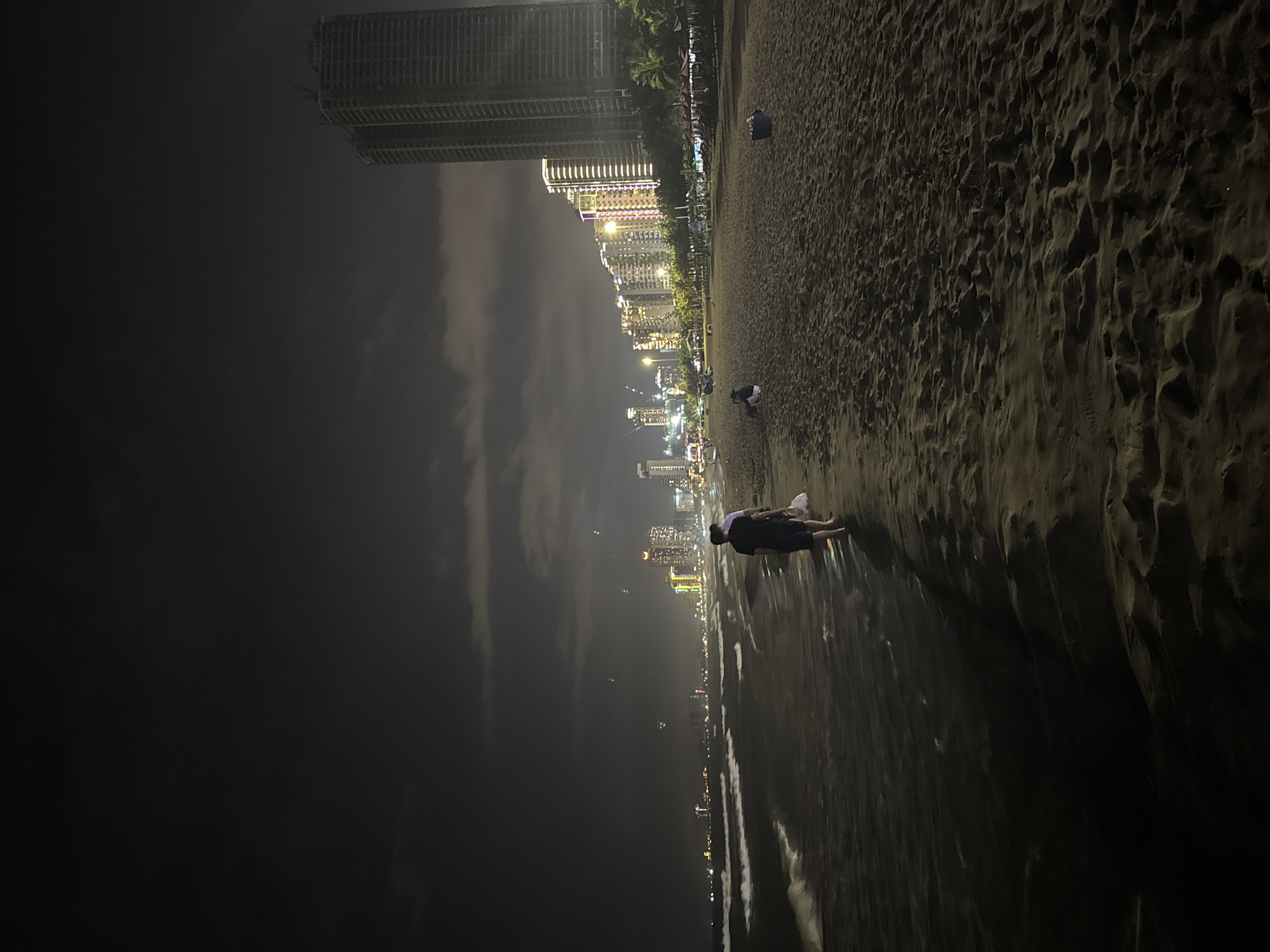 At night, a beach on the coast of Đà Nẵng, Vietnam is captured in a vertical-oriented, long shot. The scene is dimly lit, with city lights illuminating the distant skyline. The beach extends from the lower right to the upper center of the frame. The sand is dark and appears slightly wet near the water. Along the edge of the beach, there is a person lying on the sand, oriented perpendicular to the shoreline. Further up the beach, two more individuals are visible, one seated and another standing. In the background, a series of high-rise buildings lit up along the coast make up the city’s skyline. A taller, unfinished building is visible on the left. The sky is dark and partially obscured by clouds. Overall, the scene conveys a sense of quiet nighttime activity along the beach with the city as a backdrop.