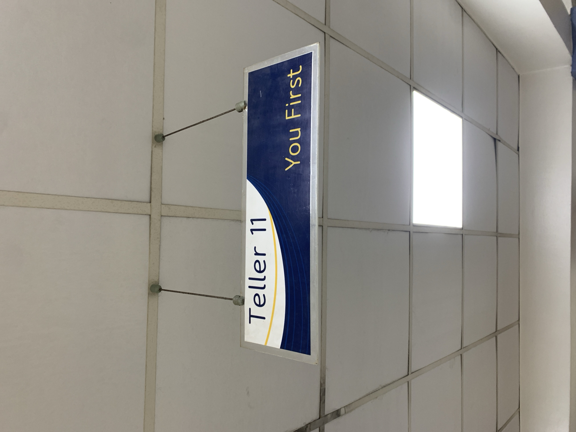 A close-up shot depicts a sign indicating "Teller 11" and "You First." The sign is vertically oriented and suspended by two thin cables from a metal grid structure against a backdrop of rectangular, white panels. The lighting is bright, with a square light panel visible in the background. The scene is sterile and institutional in appearance. The city of Ikorodu, Nigeria is not discernible from the image.