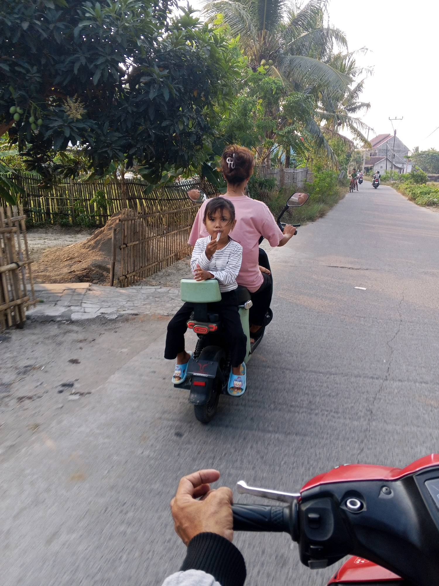 Two individuals, a child and an adult, ride on a scooter on a paved road. The child, seated behind the adult, is wearing a striped long-sleeved shirt and dark pants with blue sandals. The adult, driving the scooter, wears a pink shirt. The child holds a white object to their mouth. The scooter has a light green passenger seat and a black rear fender with a red taillight. In the background, a bamboo fence lines a sandy area with a pile of dirt. Lush green trees, including palm trees, and houses are visible. Other people on motorcycles are seen further down the road. The scene appears to be in Gosara, Indonesia, given the characteristic tropical vegetation and housing style. The image is captured from the perspective of someone riding another vehicle, as evidenced by the hand and scooter controls in the foreground. The sky is bright, suggesting daytime.