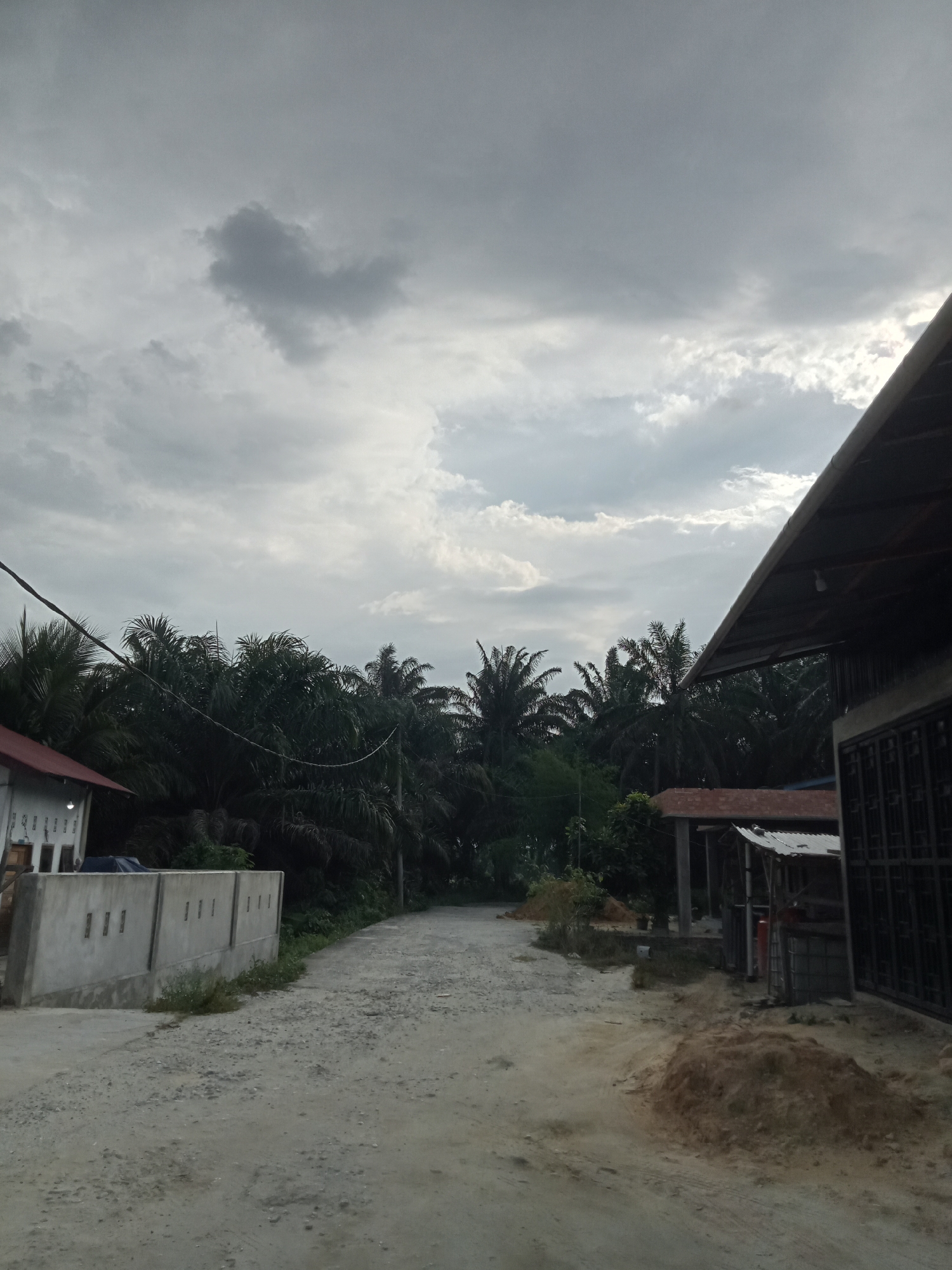 The image depicts a rural road in Melayu Besar Kota, Indonesia, with a cloudy sky overhead. On the left, a white building with a red roof is visible, next to a grey concrete wall with rectangular openings. A dirt road curves into the distance, lined by lush green palm trees. On the right side of the road, a structure with a dark-colored roof stands, partially obscuring a dark metal gate. Piles of dirt and some vegetation are present along the roadside. The overall scene suggests a residential or agricultural area with a simple, rustic atmosphere.