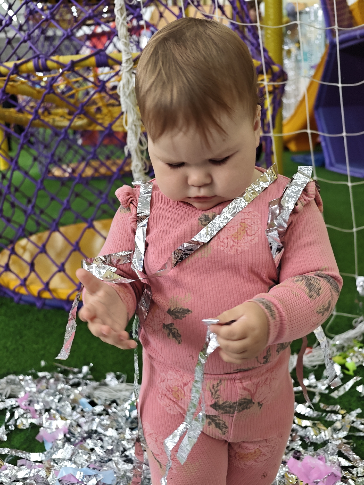 A toddler, with light brown hair, stands centered in an indoor children's play area in the city of Chaikovsky, Russia. The child is dressed in a pink, ribbed long-sleeved top with ruffle detailing on the shoulders and matching pants, both featuring a subtle floral pattern in darker pink and brown tones. Numerous strands of crinkled silver metallic foil are draped over the child's shoulders and torso. The child is actively manipulating a piece of this silver foil in their right hand, gazing downwards with a focused expression. Their left hand is open and slightly extended forward. The child stands on a green surface resembling artificial turf, which is densely covered with scattered pieces of silver, light pink, and light blue foil confetti. In the background to the left, a play structure featuring dense purple netting stretched over yellow support bars is visible. To the right, a white net, consistent with a safety barrier or goal net, is present, behind which blurry colorful play equipment, including a purple slide-like structure, is discernible.