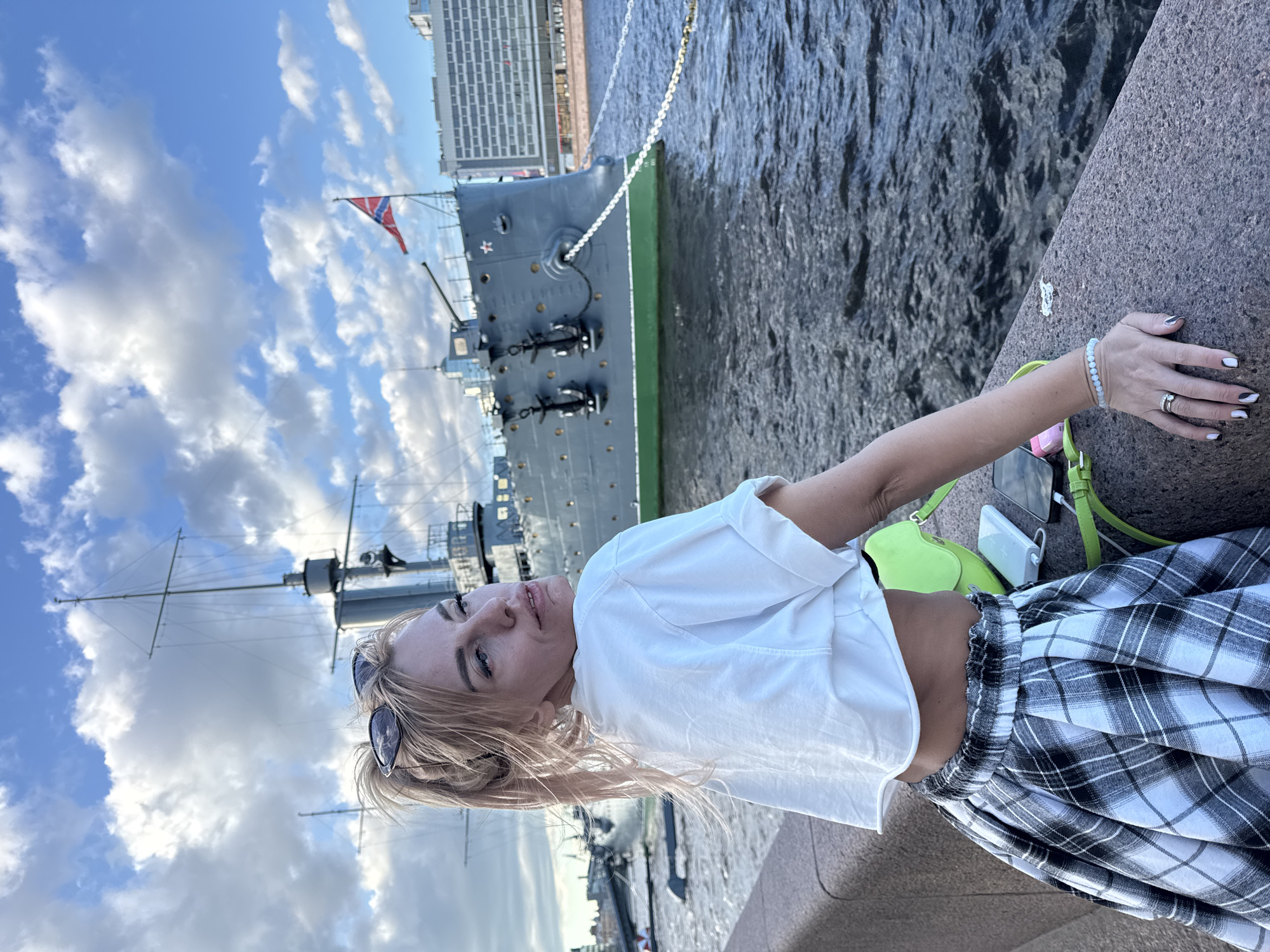 A young Caucasian woman with blonde hair tied in a ponytail and sunglasses perched on her head is standing next to a stone embankment. She is wearing a white crop top and a black and white plaid skirt. Her left arm is resting on the embankment, and her right hand is extended towards the viewer. Behind her, a large gray ship with a green hull is docked in the water. A Russian flag is flying from the ship's mast. The sky is blue with scattered white clouds. The scene appears to be along the Neva River in Saint Petersburg, Russia, with the Aurora cruiser visible in the background.