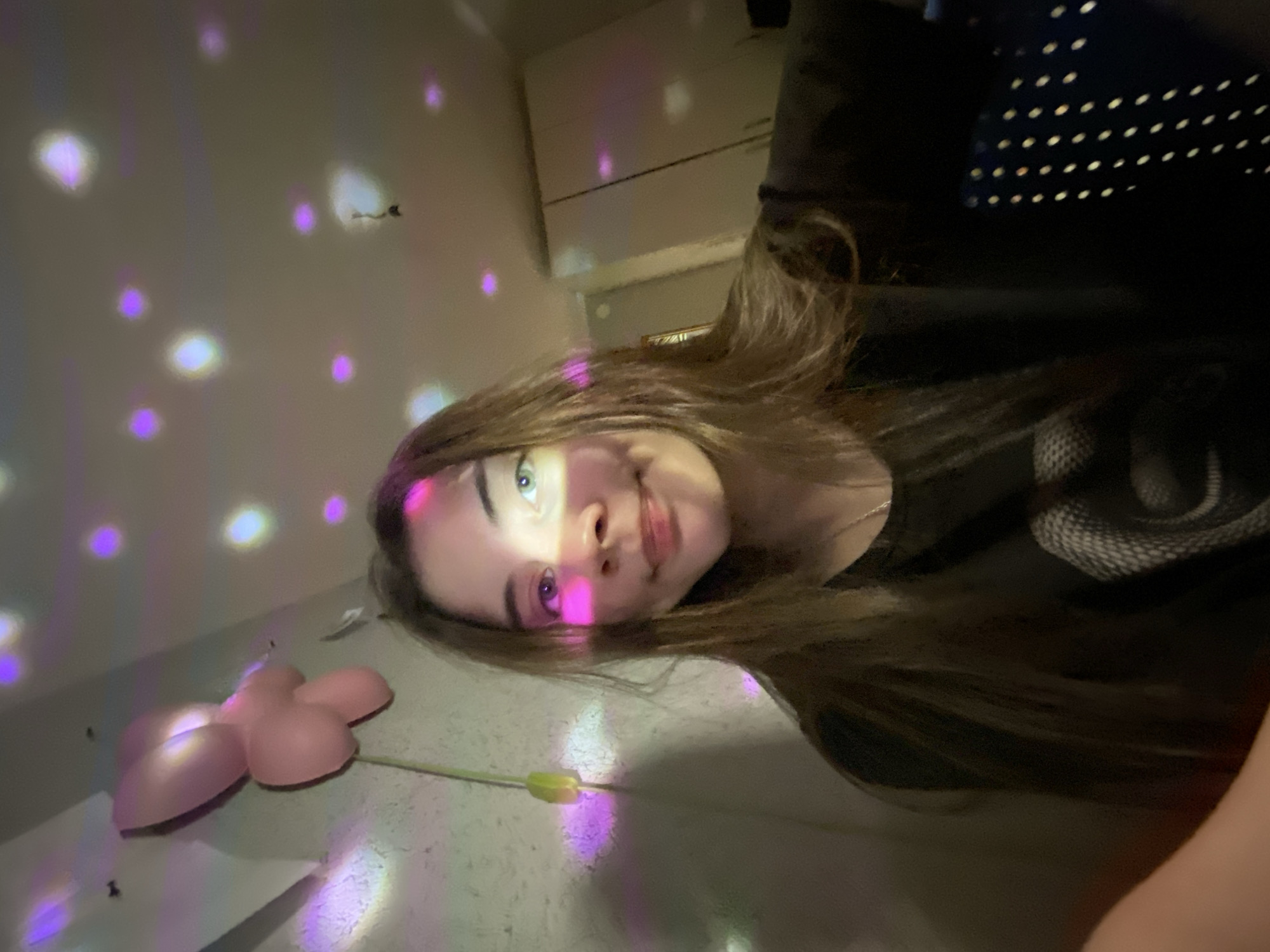 The image is a selfie of a young woman with long brown hair. She is wearing a black shirt, and part of a black object with white dots is visible in the upper right corner. Her face is illuminated by purple and pink light spots. A pink, flower-shaped object is visible on the wall. There are also indistinct objects visible in the background. There is no information available from the image to confirm or deny that the location is in the city of Votkinsk, Russia.