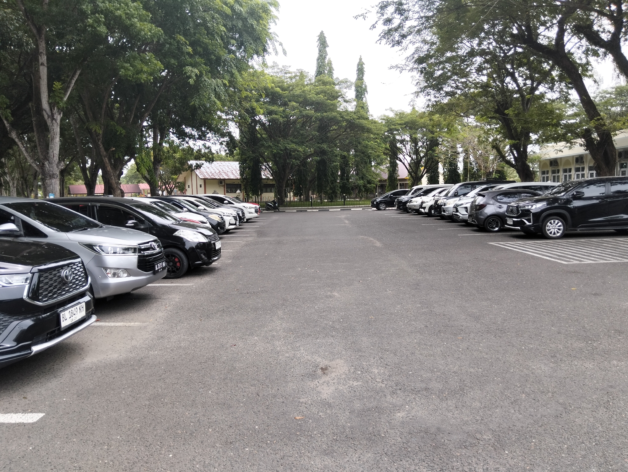 The image shows a parked row of cars in a lot with mature trees overhead and a building in the background. The cars are of various colors and models, with license plates visible on some of them. The parking lot is paved with asphalt, and there are white painted lines demarcating the parking spaces. The trees are large and leafy, providing ample shade. The building appears to be a large institutional structure with multiple windows. The scene is calm and orderly, suggesting a typical day in this location. The presence of parked cars and the building indicate a functional area within the city. The overall atmosphere is one of serenity, with the natural elements of the trees complementing the man-made structures.