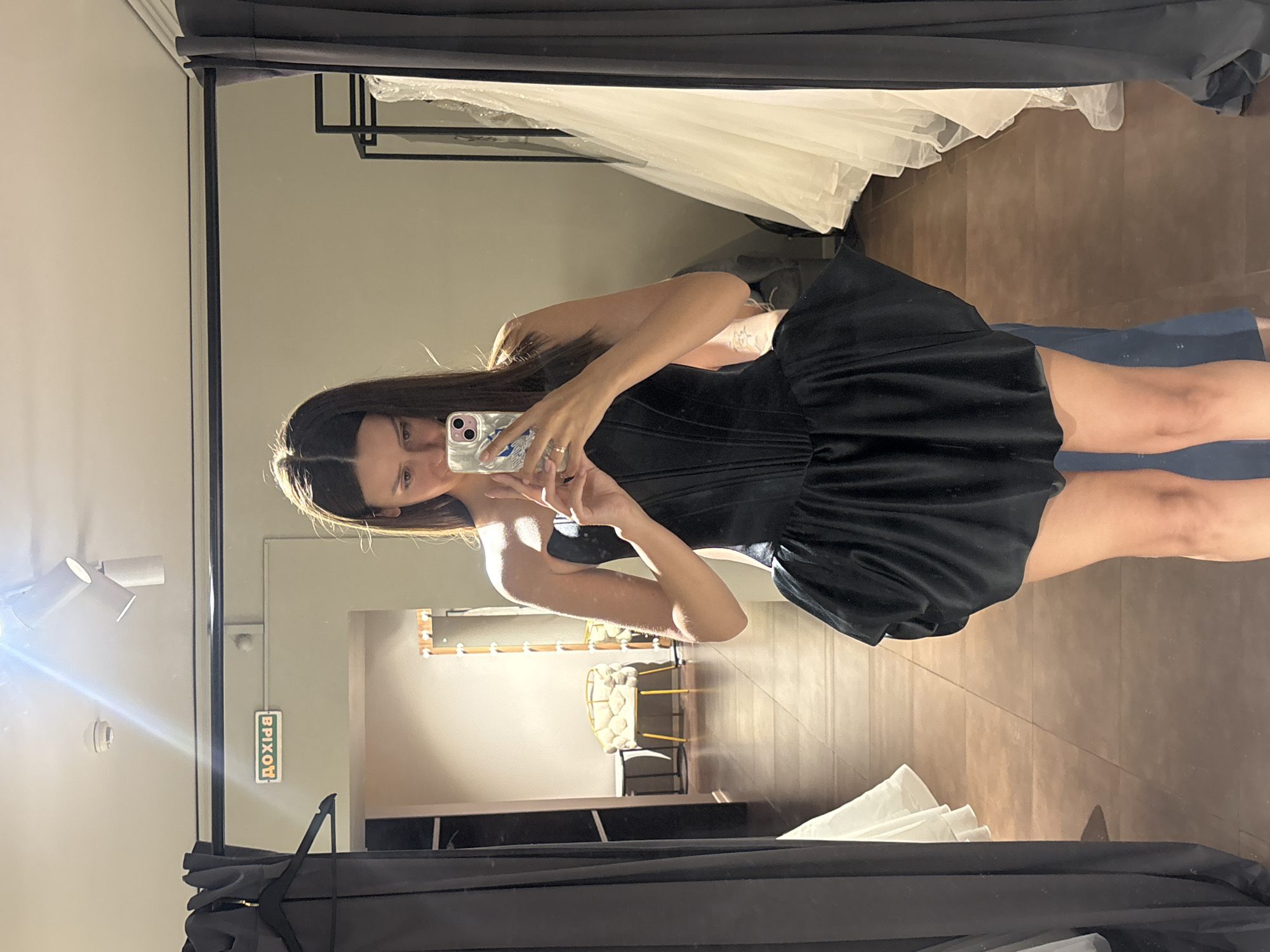 A young woman is taking a mirror selfie in a fitting room. She is wearing a black strapless dress with a structured bodice and a voluminous, flared skirt. Her hair is long and dark, and she is holding a smartphone in her right hand. The fitting room appears to be in a clothing store, with racks of dresses visible in the background, including several white gowns that resemble wedding dresses. There is also a chair with a distinctive design, and a sign on the wall that appears to be in Cyrillic. The lighting suggests an indoor setting, likely a retail environment. The image does not provide any direct information about the city of Krasnoyarsk, Russia.