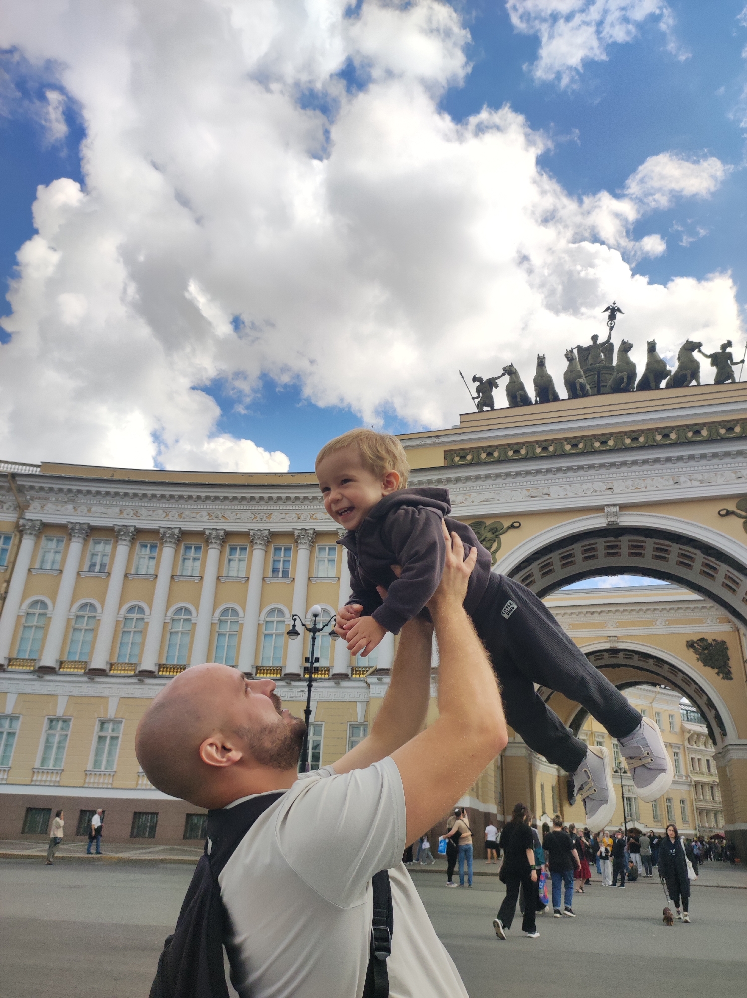 In this low-angle shot, a bald man is throwing a young child into the air. The man is wearing a grey t-shirt and a backpack. The child, dressed in a dark grey tracksuit and white sneakers, is laughing. In the background, a grand yellow building with white columns and arched gateways dominates the scene, adorned with statues of horses and chariots on its rooftop. The sky above is partly cloudy with a bright blue hue. The location is the General Staff Building Arch, a prominent landmark in Palace Square, Saint Petersburg, Russia.
