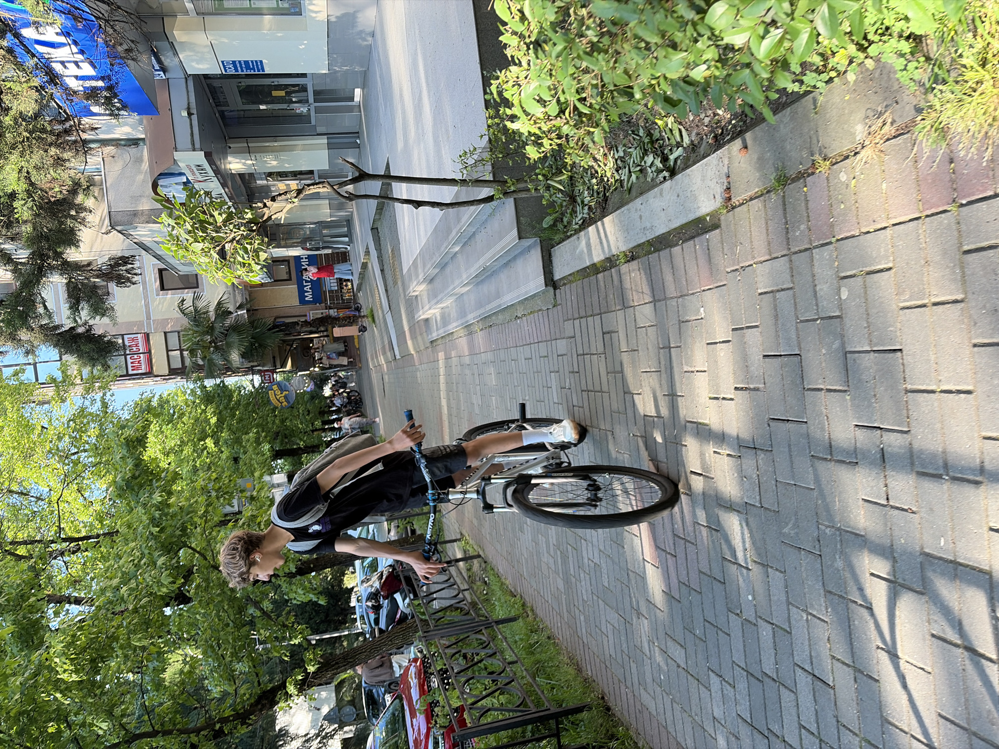 The image depicts an outdoor street scene in the city of Sochi, Russia, during daytime, characterized by bright sunlight and long shadows indicating morning or late afternoon. The foreground features a sidewalk paved with grey rectangular interlocking pavers laid in a herringbone pattern. Small weeds are visible in the pavement cracks and along the curb.
The primary subject is a young male, appearing to be a teenager, with light brown wavy hair, wearing a black t-shirt, dark shorts, white socks, and white sneakers. He carries a grey backpack and wears white in-ear headphones. He is actively riding a silver-framed bicycle with front suspension, black tires, and blue accents on the handlebars, where "NITRO" is visible on the stem. His body is oriented towards the right side of the frame, but he is looking over his left shoulder. His right foot is on a pedal, and his left foot is either touching the ground or off it.
To the left of the sidewalk, a dark metal fence separates the pedestrian area from a strip of green grass and foliage. Behind this, several parked vehicles are visible, including a red SUV, dark sedans, and a scooter or motorcycle. Mature trees with dense green leaves line this side of the street, casting significant shadows onto the pavement.
To the right of the sidewalk, a raised concrete area features two steps leading up to the entrance of a commercial building. A small, sparsely leafed tree grows in a bed adjacent to these steps, next to a planter box filled with green bushes. The multi-story buildings on the right side display various commercial signs in Cyrillic script: a prominent blue awning reads "АПТЕКА" (Pharmacy), another sign indicates "КУХНИ" (Kitchens), and further signs show "МАГАЗИН" (Shop), "ОСАГО" (OSAGO - Compulsory Motor Third Party Liability Insurance), and "МАС САЖ" (Massage) on an upper floor. Palm trees are also visible near these buildings.
In the background, the street continues with more buildings and trees, and several other pedestrians are present in the distance. The sky is clear and bright blue.