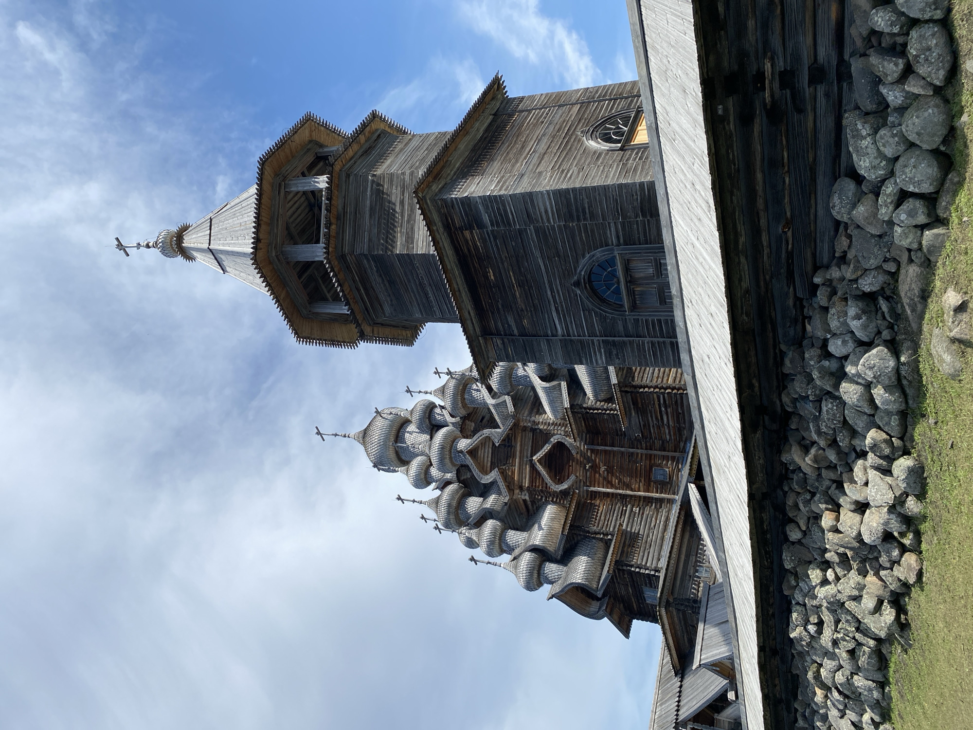 The image shows the Church of the Transfiguration, a wooden architectural monument in the city of Boyarshchina, Russia. The church is a prominent structure with multiple domes and a tall bell tower. It is made of weathered wood, giving it a rustic and historic appearance. A stone wall is visible in the foreground, with grass surrounding it. The sky is blue with scattered clouds. There are no people visible in the image.