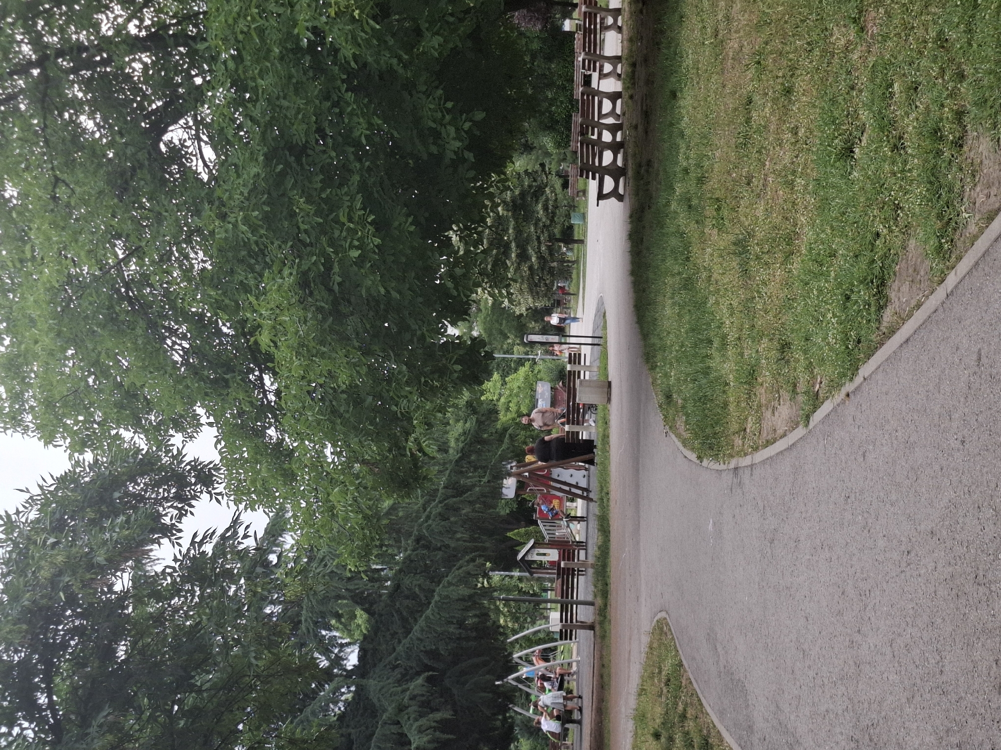 In a park setting, a paved pathway curves through grassy areas and under the shade of large trees. To the left, a playground is visible with swings, slides, and climbing structures, where several children and adults are present. Benches are situated along the pathway. To the right, more benches are lined up. In the background, other park visitors can be seen walking. The sky is overcast.