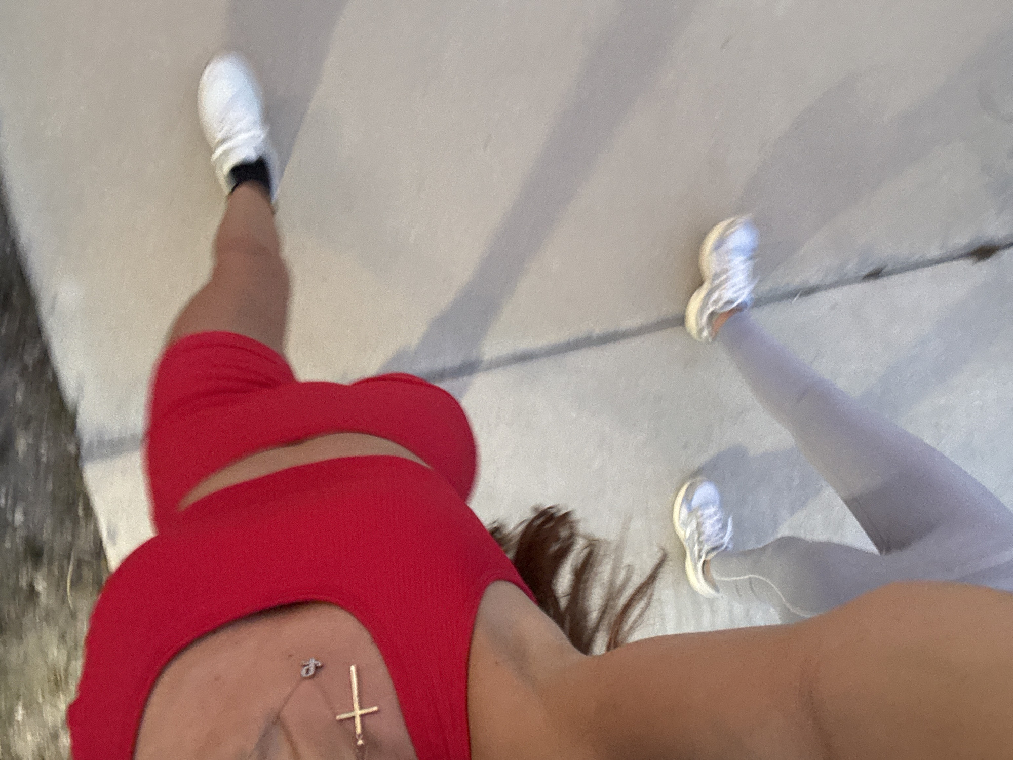 An individual is captured in a dynamic, low-angle shot, appearing to be in motion outdoors. They are dressed in a vibrant red athletic top and light gray leggings, complemented by white athletic shoes and black socks. A silver cross necklace is visible on their chest, resting on the red top. The background consists of a textured, light-colored surface, possibly a sidewalk or pavement, with distinct shadows cast, indicating an outdoor environment under bright lighting. The image is slightly blurred, suggesting movement.