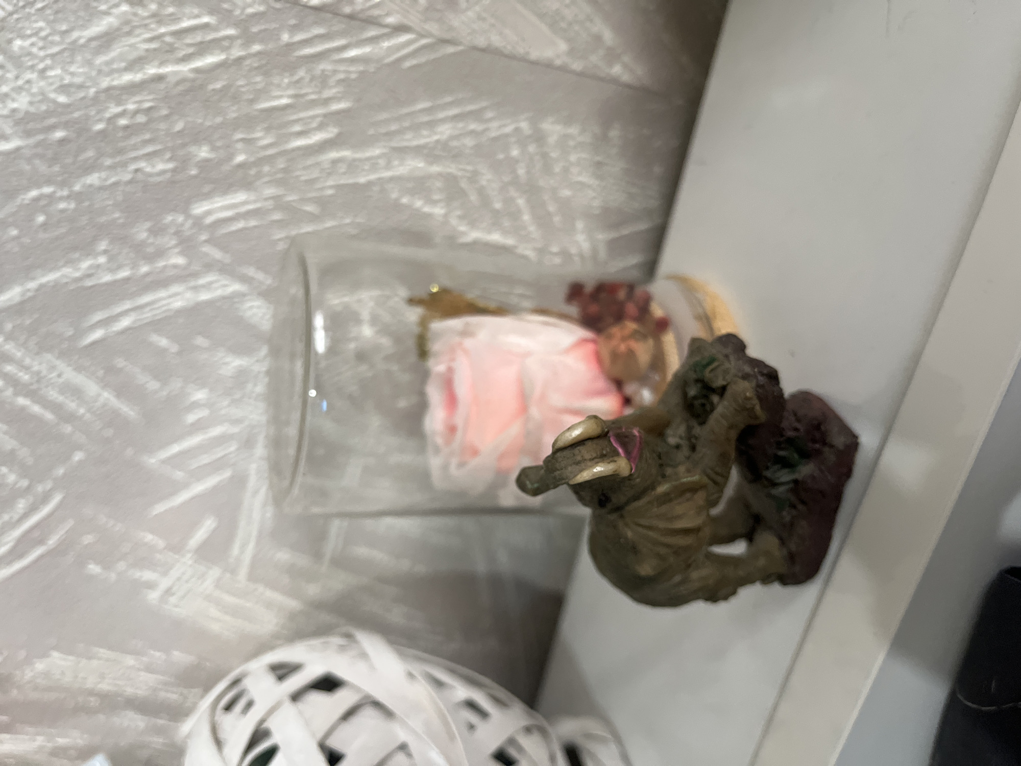 The image depicts a still life on a white shelf. A glass jar containing a pink flower and dried red elements lies horizontally on the shelf. Next to the jar is a decorative figurine of a monkey or ape, appearing to be resting on or interacting with a dark, possibly rocky base. Below the jar, partially visible, is a white, round, lattice-like object. Part of a black object is visible on the right side of the image. The location in the city of Sofia, Bulgaria is not evident from the image.