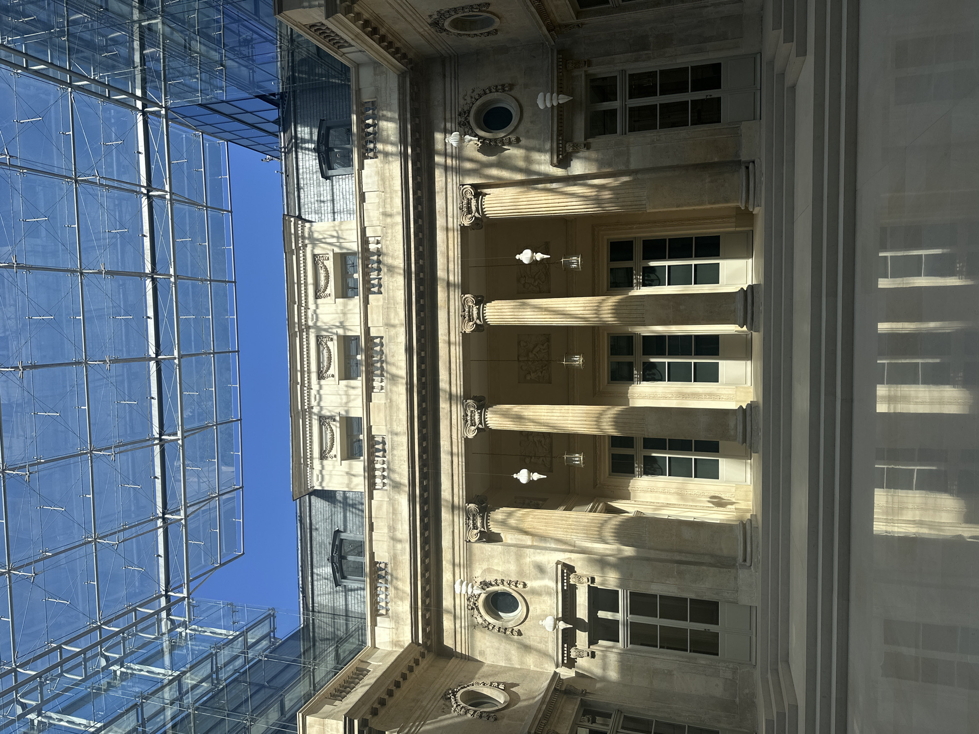 Looking up at a classical building with ornate details, columns, windows, and decorative elements. To the left, a modern glass building with a grid-like structure. Blue sky is visible between the two buildings. The location is likely Paris, France, considering the architectural style. No people are visible.