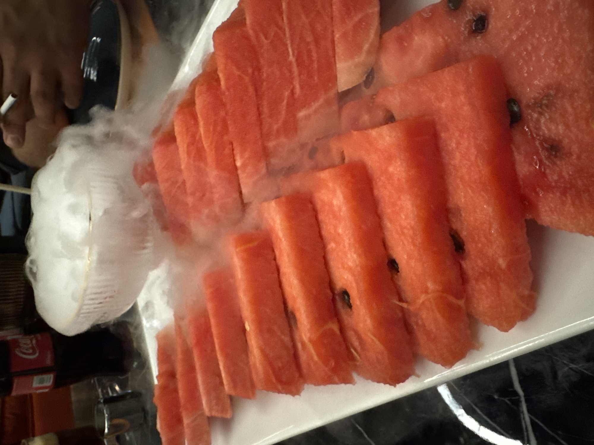 A high-angle shot presents a platter of sliced watermelon. The watermelon slices are arranged in a row on a white rectangular plate. Some of the slices have visible black seeds. Around the watermelon, there is a cloud of white vapor, likely created with dry ice for a theatrical effect. A person's hand is visible in the background, holding what appears to be a thin, white cigarette. Behind the platter, partially visible, is a can of Coca-Cola. The surface the platter is placed on appears to be black with white marbling. There is insufficient information to confirm the location in Yerevan, Armenia.