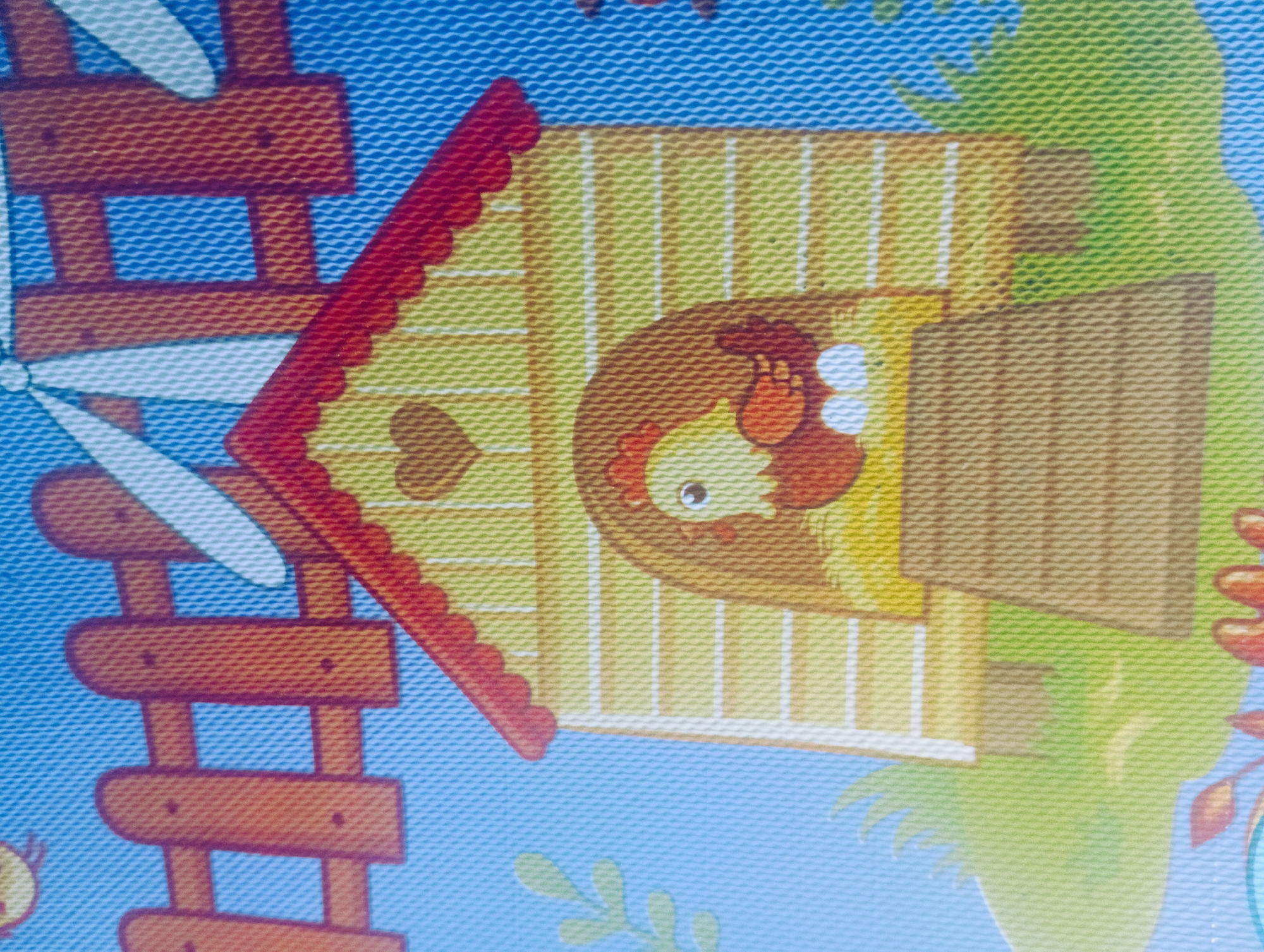 This is a close-up, top-down, medium shot of a cartoon chicken coop and fence depicted on a blue mat with a textured surface. The chicken coop is yellow with a red, scalloped roof and a brown wooden ramp leading into it. A cartoon chicken with a red comb and wattle is visible inside the coop, sitting on a nest with two white eggs. To the left of the coop is a section of orange wooden fence. In the background, there are white windmill blades against a blue sky. Green grassy mounds are at the base of the coop, and a few stylized green leaves are to the left. The image appears to be a detail from a play mat, possibly for children. There is no indication of the location in Kutasari, Indonesia.