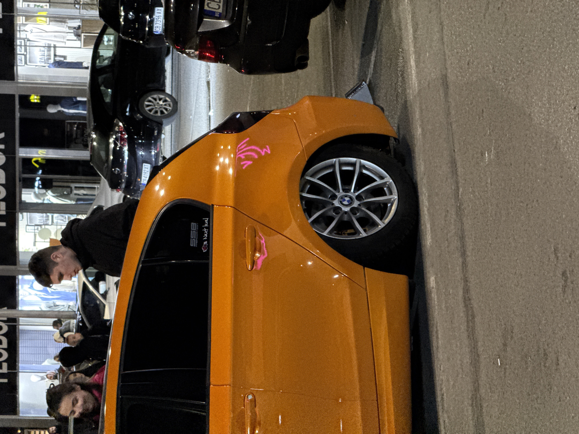 The image depicts an orange BMW parked on a street, likely in Sofia, Bulgaria, given the visible license plates and store signs. A group of people, including a man leaning into the car, is gathered around the vehicle. Other cars are parked nearby. The scene suggests a casual street gathering or an interaction related to the vehicle. The setting appears to be at night with adequate street lighting.