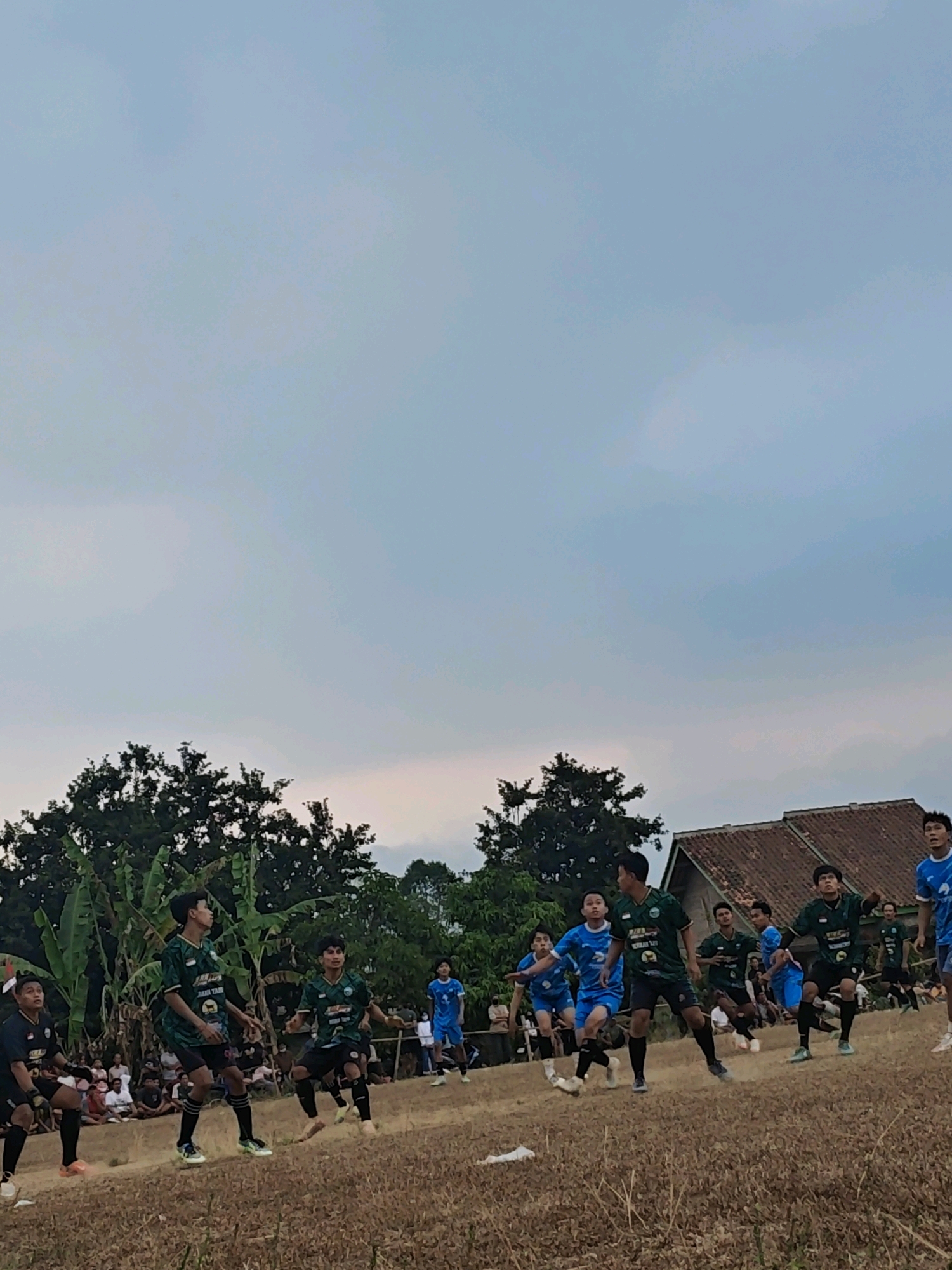 In Karang Sari, Indonesia, a group of young men in athletic uniforms are engaged in a soccer match on a dry, grass-covered field. The sky above is overcast with a pale blue and grey hue. In the background, a cluster of trees and a red-tiled roof building are visible. The players, dressed in green and blue jerseys and black shorts, are in various stages of activity, with some appearing to be in mid-action, possibly kicking or jumping. A small crowd of spectators can be seen gathered behind a low barrier in the distance. The overall atmosphere suggests a casual, local sporting event.