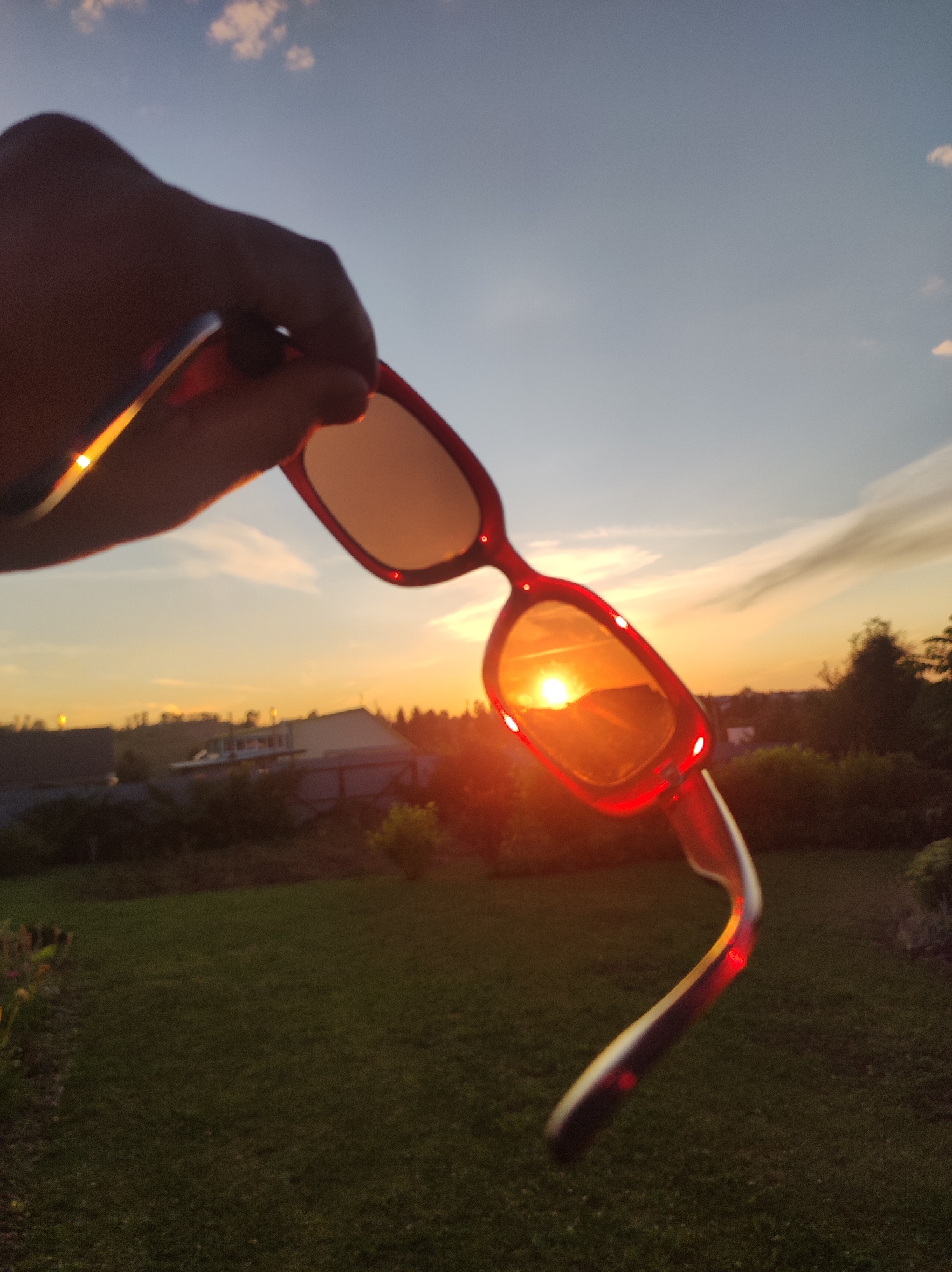 An over-the-shoulder, low-angle, medium shot shows a hand holding a pair of red-framed sunglasses in front of a sunset over a green, grassy backyard in Foky, Russia. The sunglasses are positioned so that the sun is reflected in the right lens, appearing as a bright, orange orb. The sky is a gradient of pale blue and orange, with wispy clouds. In the background, trees and rooftops are visible, silhouetted against the setting sun. The grass in the foreground is dark green, with a patch of flowers on the left side of the frame. The image is sharp and clear, with a shallow depth of field that blurs the background slightly.
